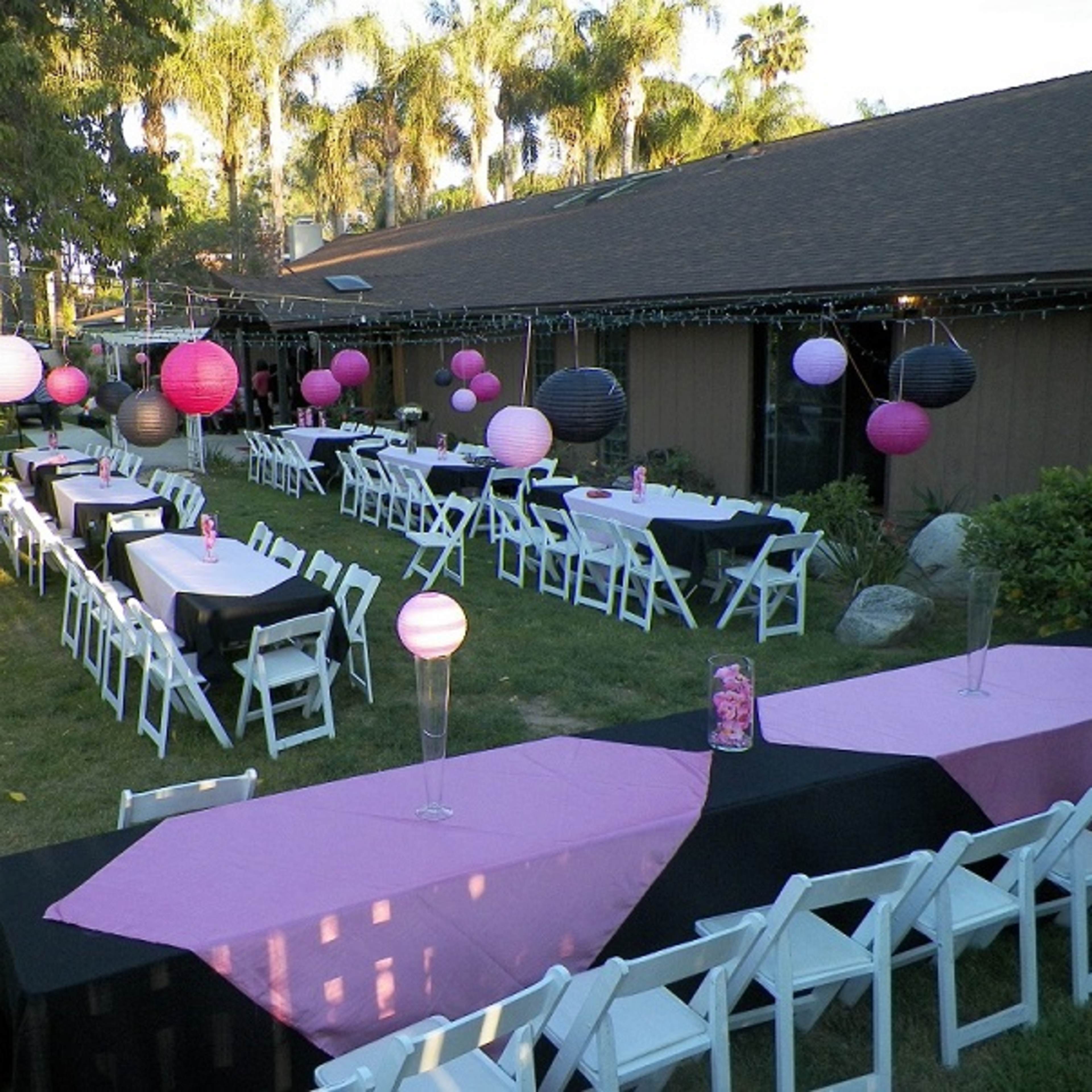 A backyard area set up for a party, featuring several long tables with black and pink tablecloths, and colorful lanterns hanging above.