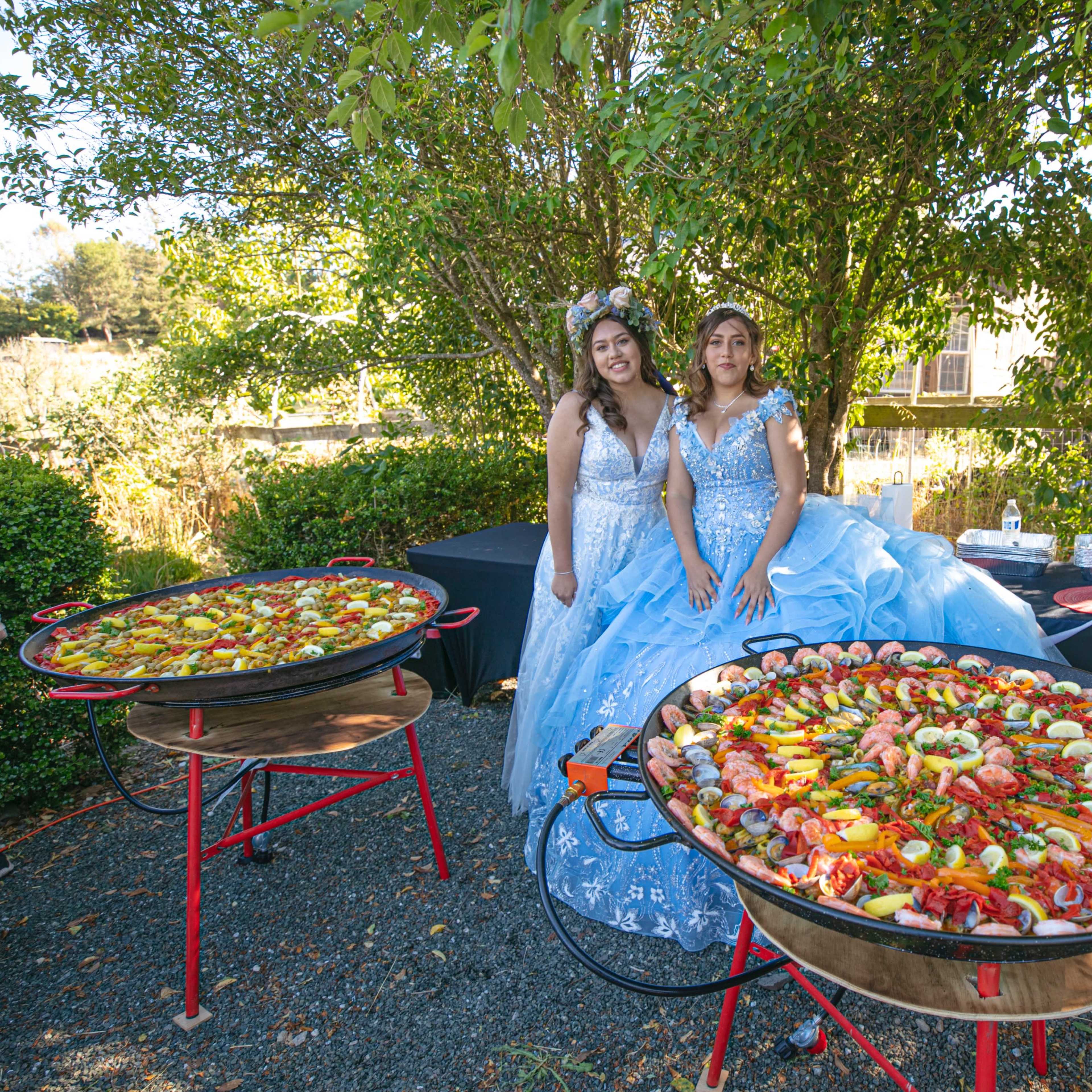 Two women in blue dresses stand beside large pans of colorful paella under a tree at an outdoor event.