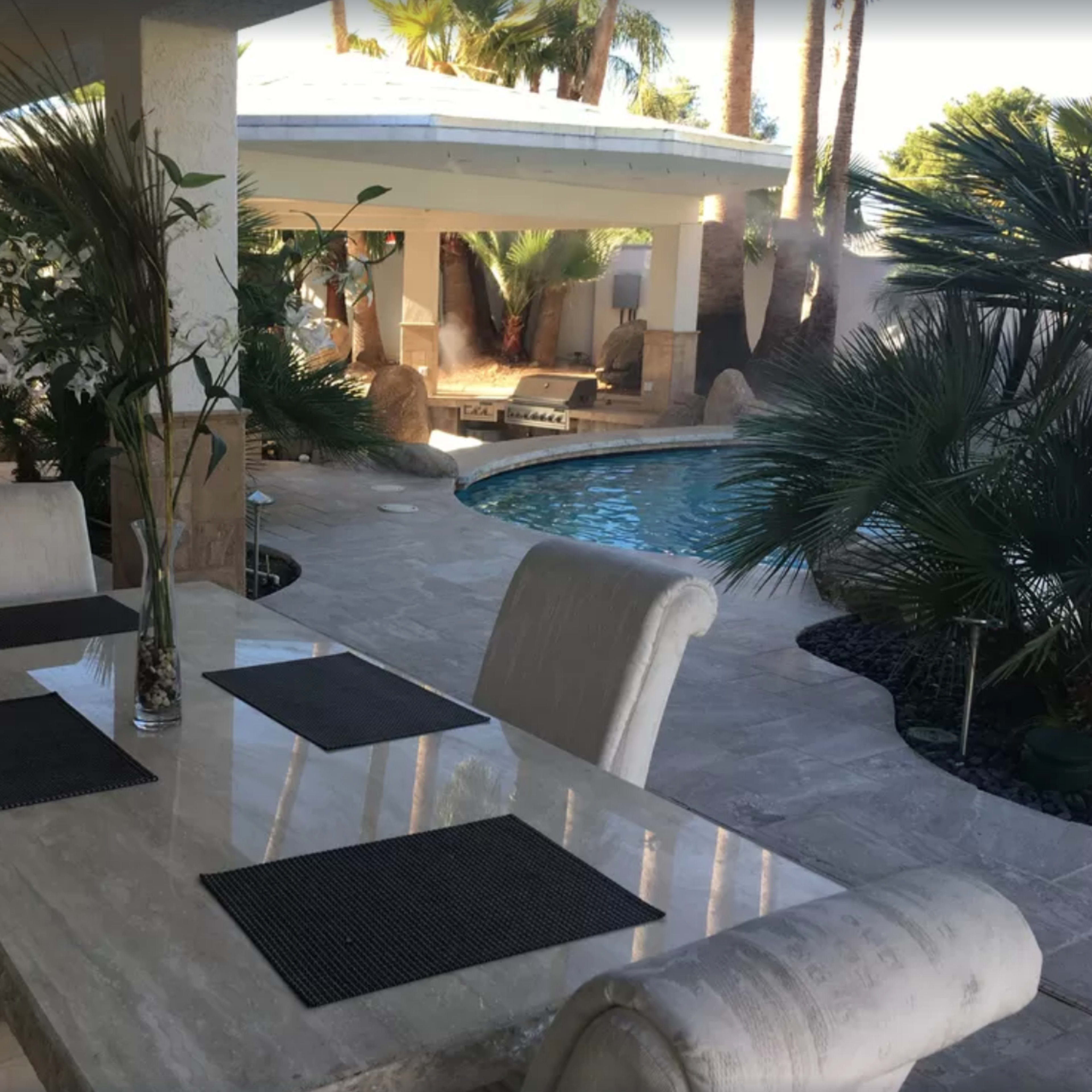 A dining table is set with placemats in a covered patio area next to a pool surrounded by palm trees.