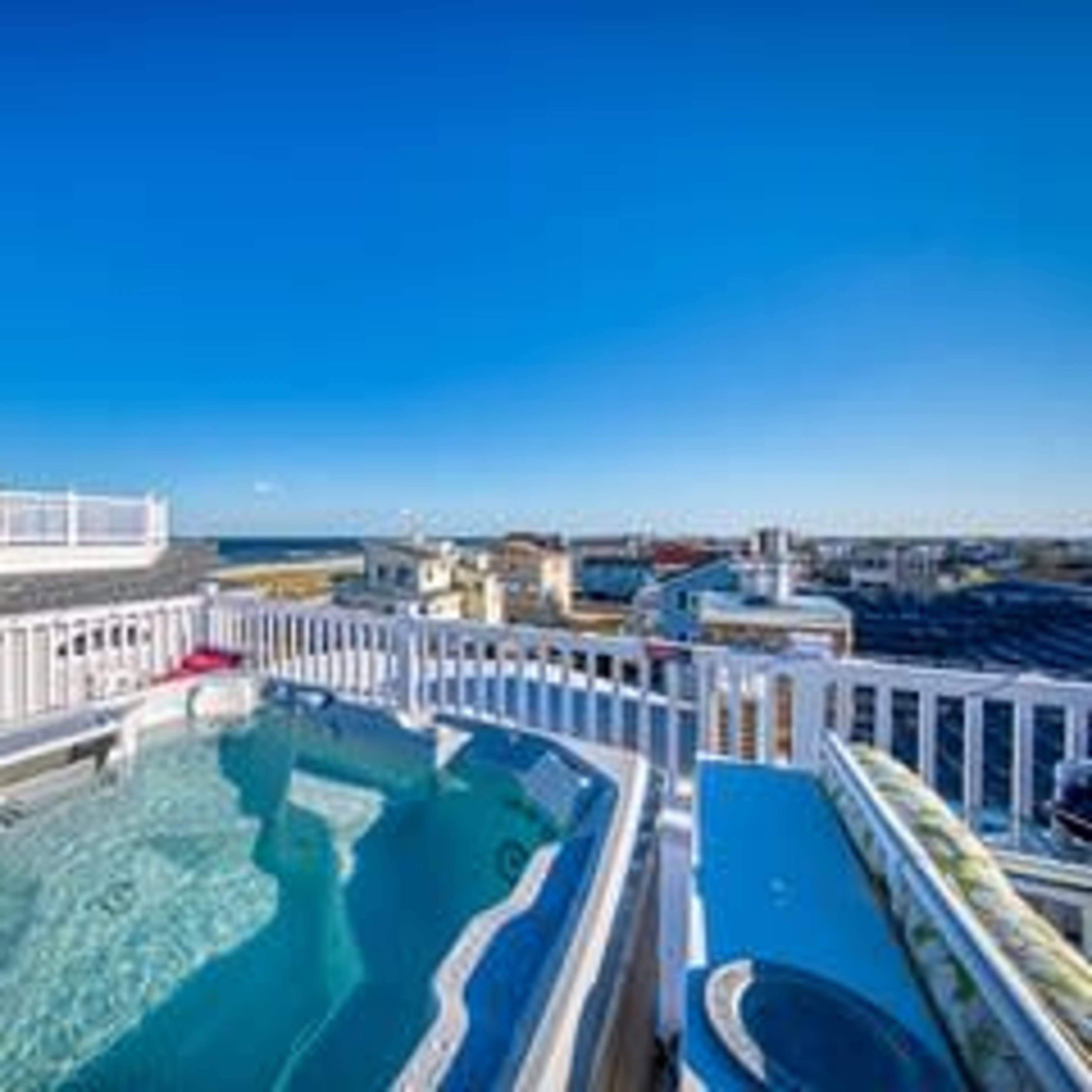 A rooftop deck features a hot tub with a view of the surrounding buildings and ocean under a clear blue sky.