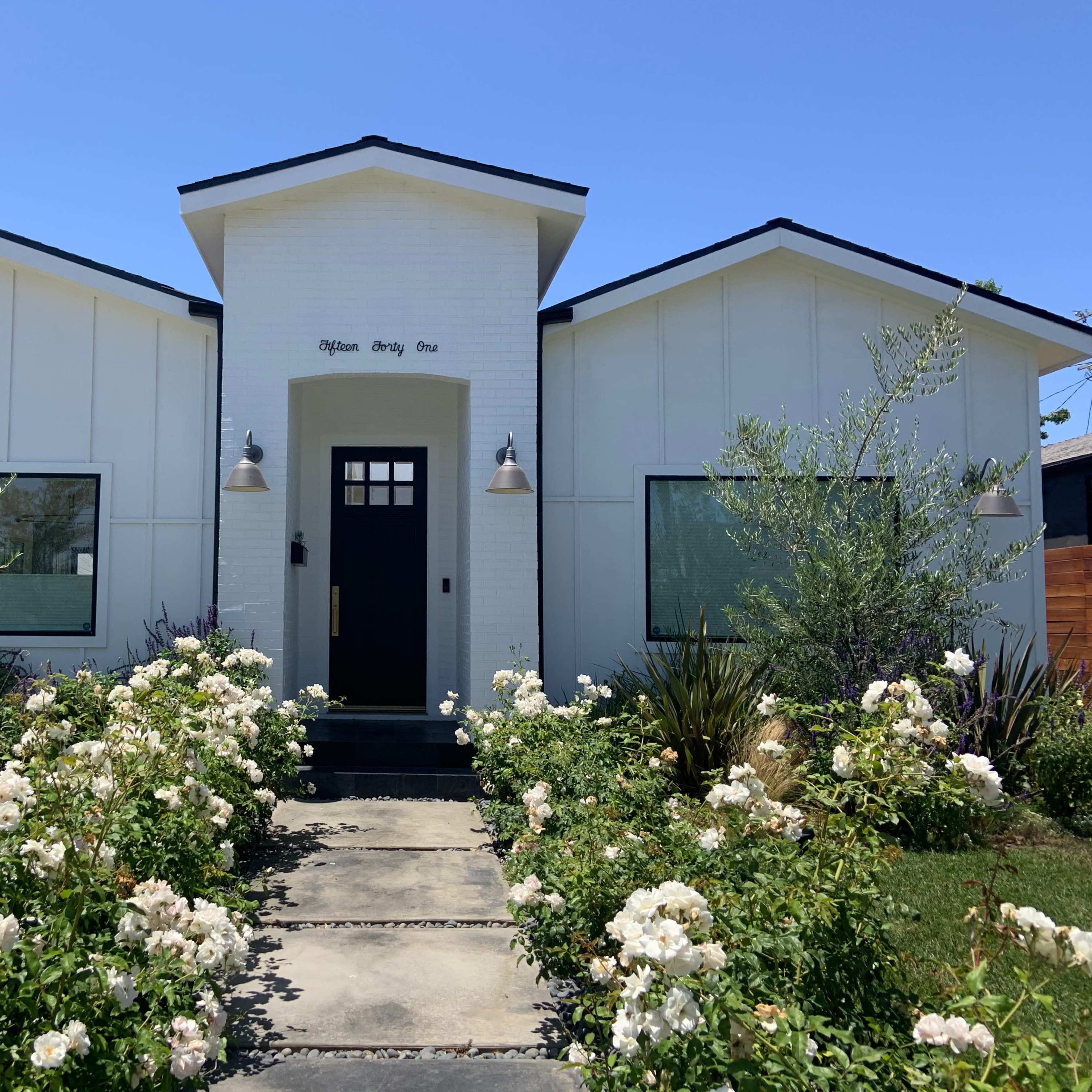 A modern white house with a geometric facade, flanked by flowering shrubs and plants, features a black front door and large windows.
