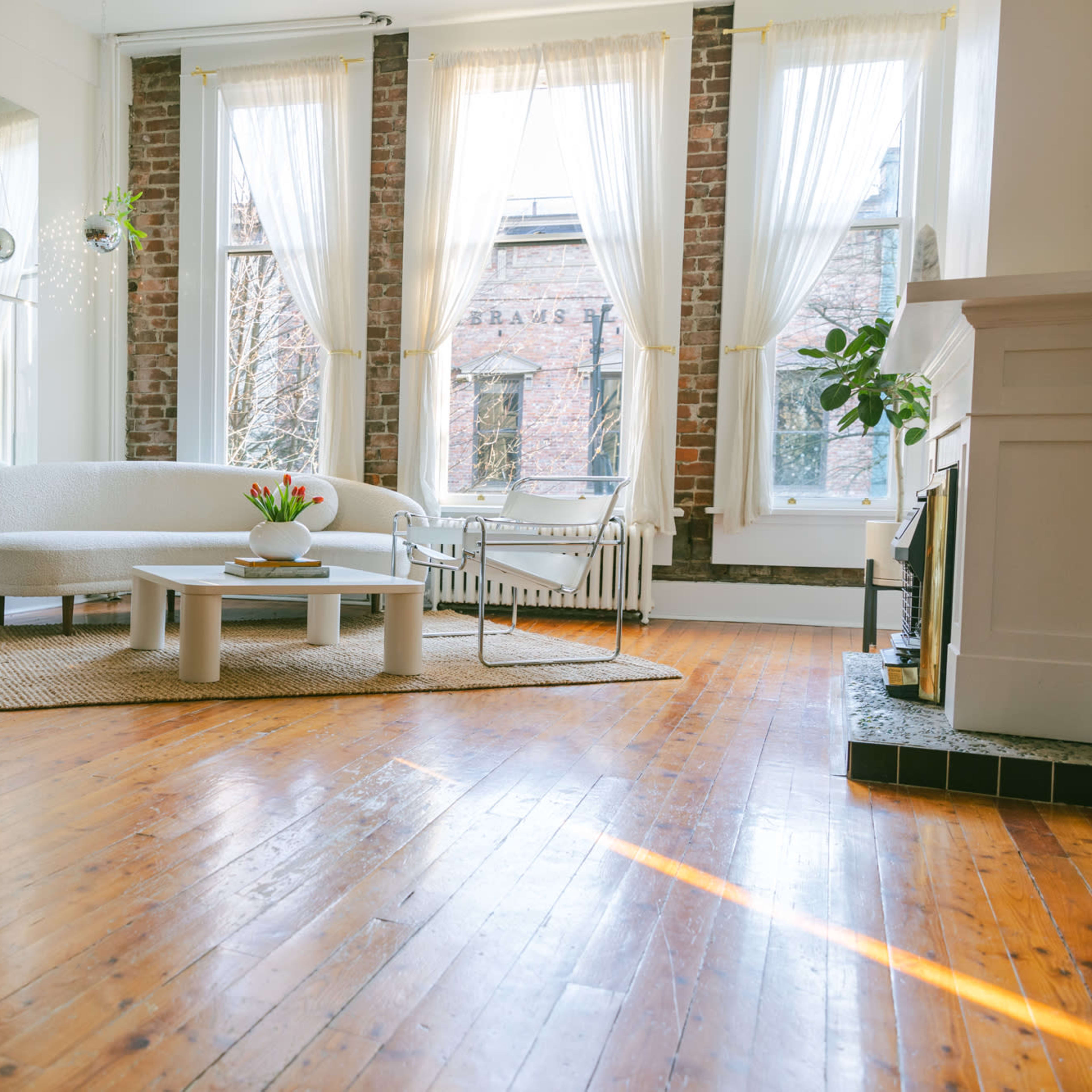 A bright living room features a curved white sofa, a round coffee table, and large windows with sheer curtains, all set on polished wooden floors.