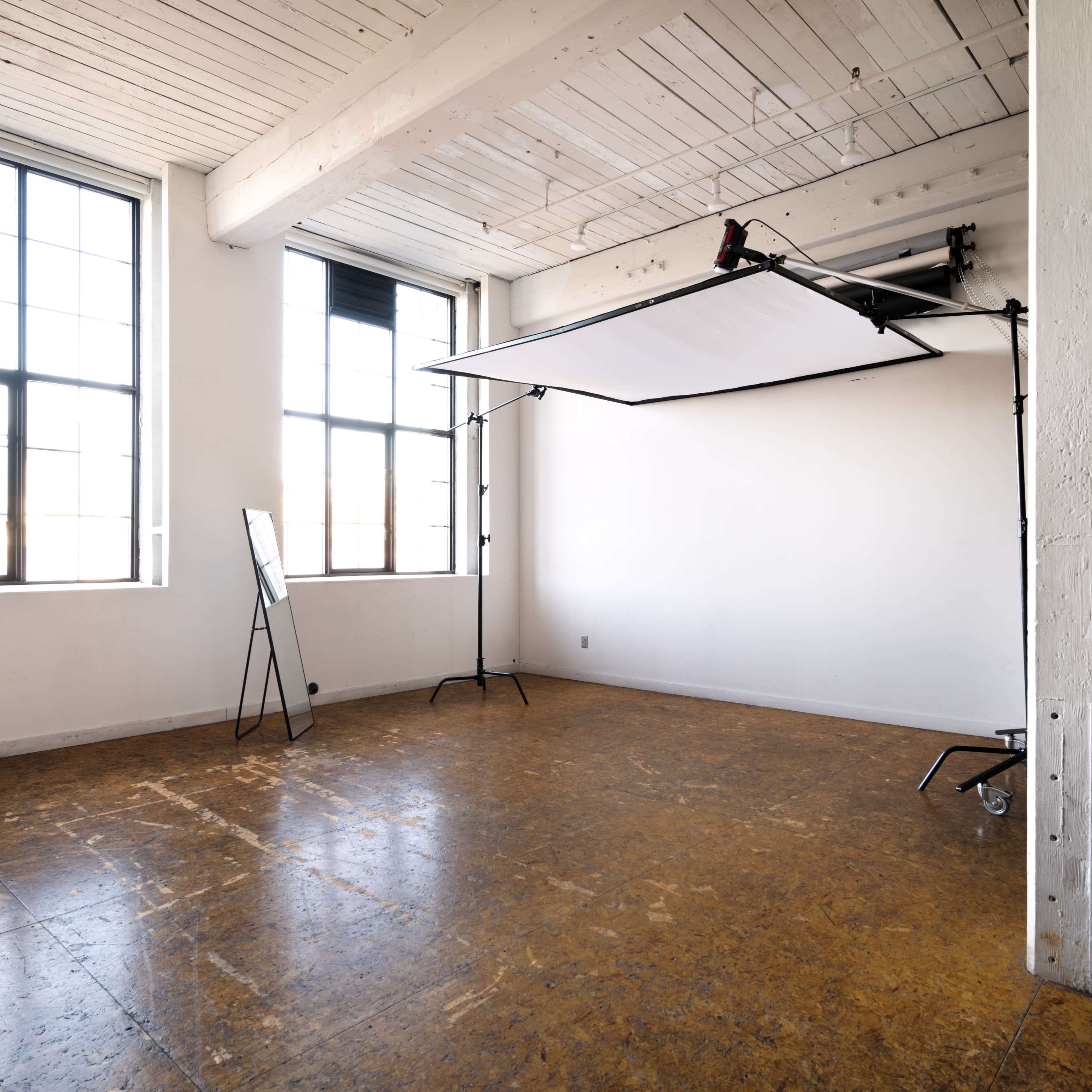 A vacant studio space with large windows, a white backdrop supported by a stand, and polished flooring.