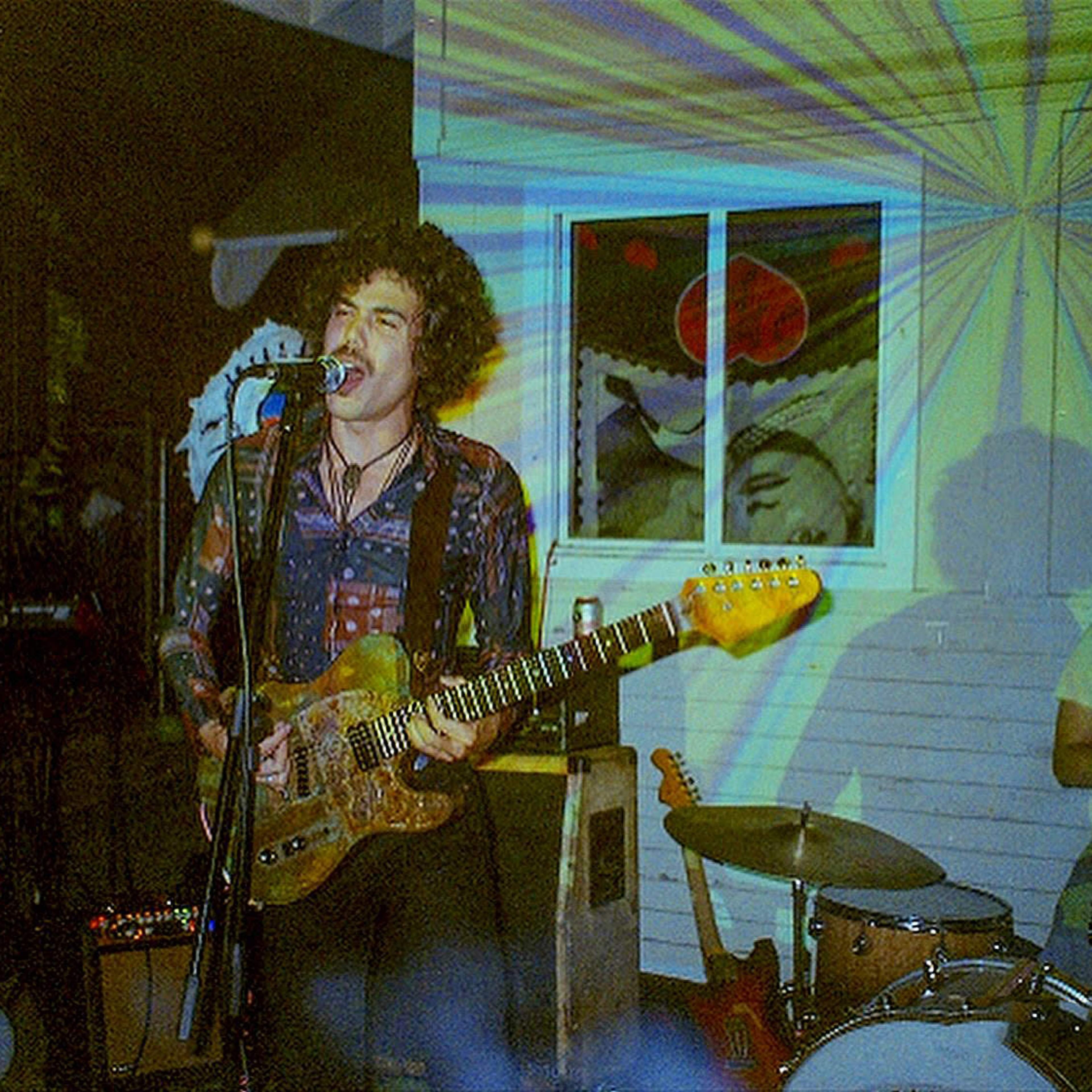 A musician plays guitar and sings on stage while a drummer sits behind a vintage drum kit in a dimly lit venue with colorful lights projected on the walls.