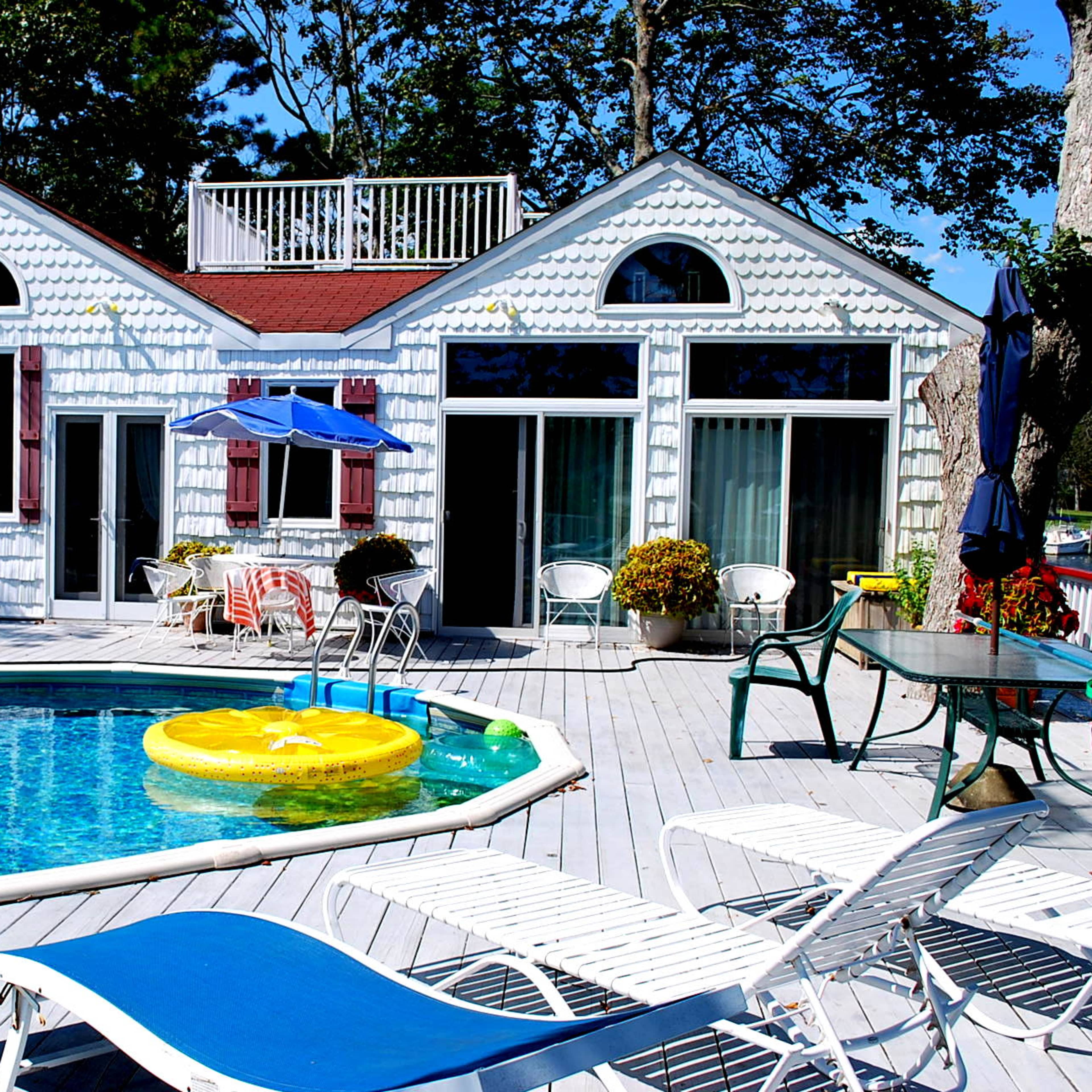A private pool area features a blue pool surrounded by lounge chairs and a white house with red accents and large windows.