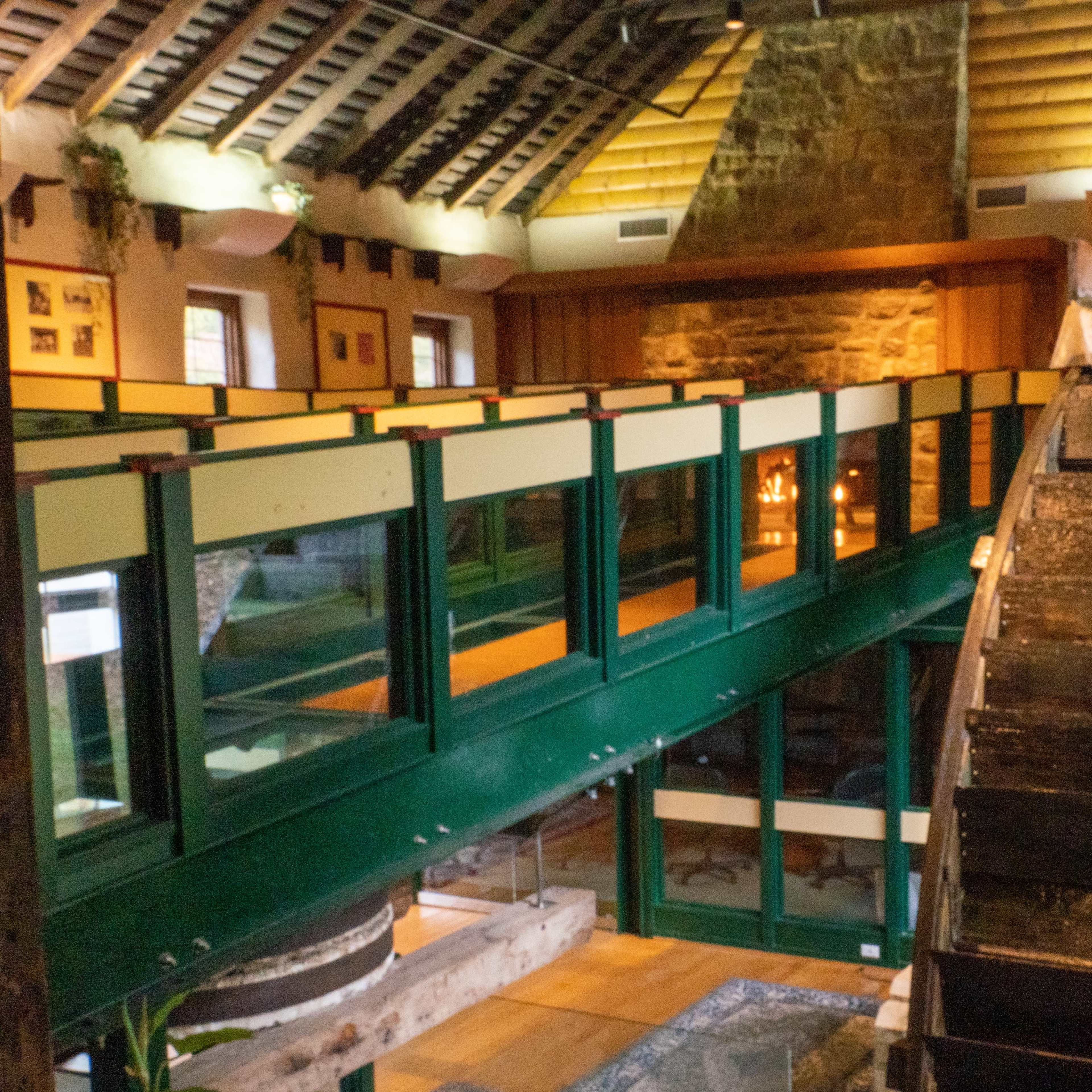 A rustic interior of a building with exposed wooden beams, featuring a large water wheel and a balcony overlooking the main space.