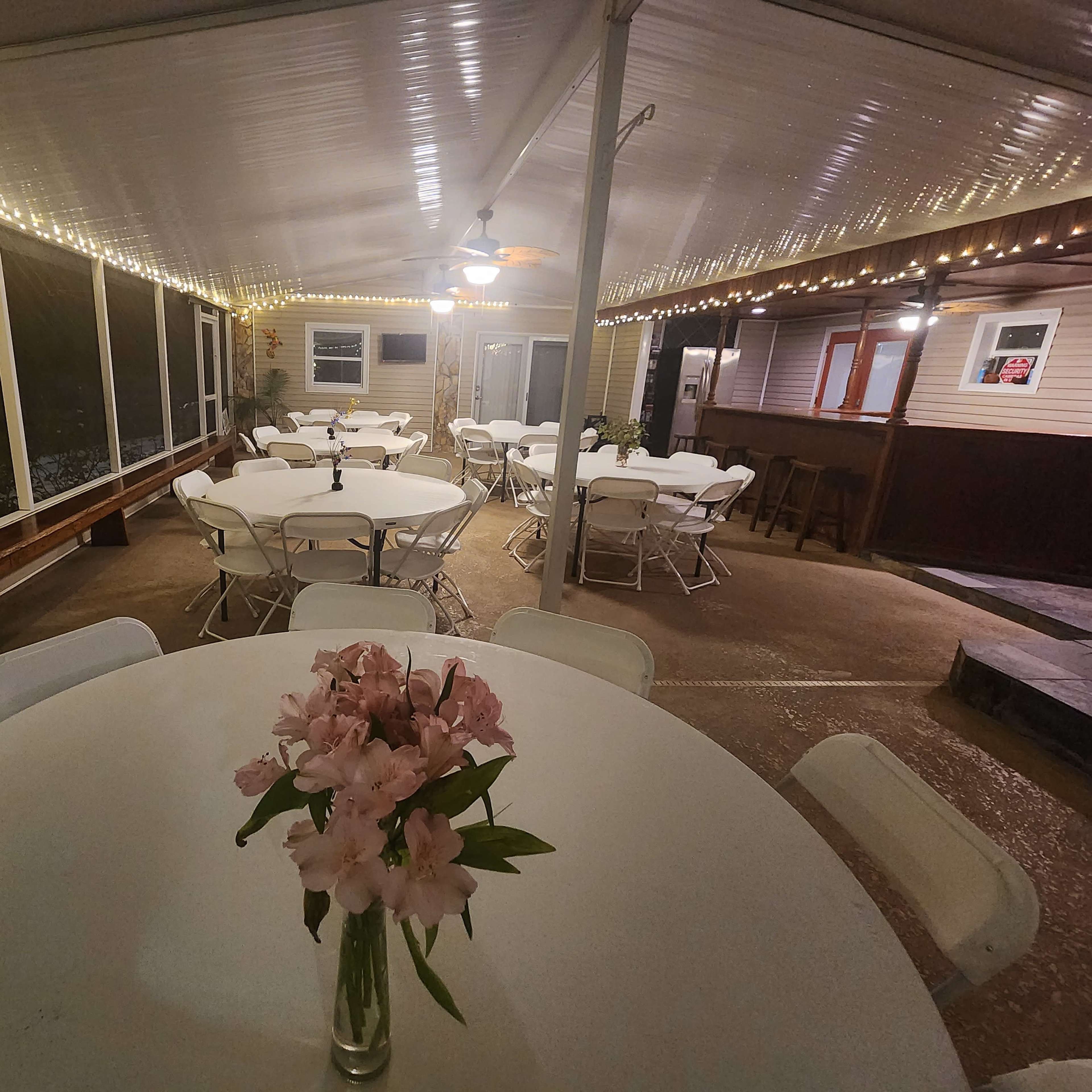 A spacious outdoor event area features round tables with white tablecloths, surrounded by string lights and a decorative vase of pink flowers on a central table.
