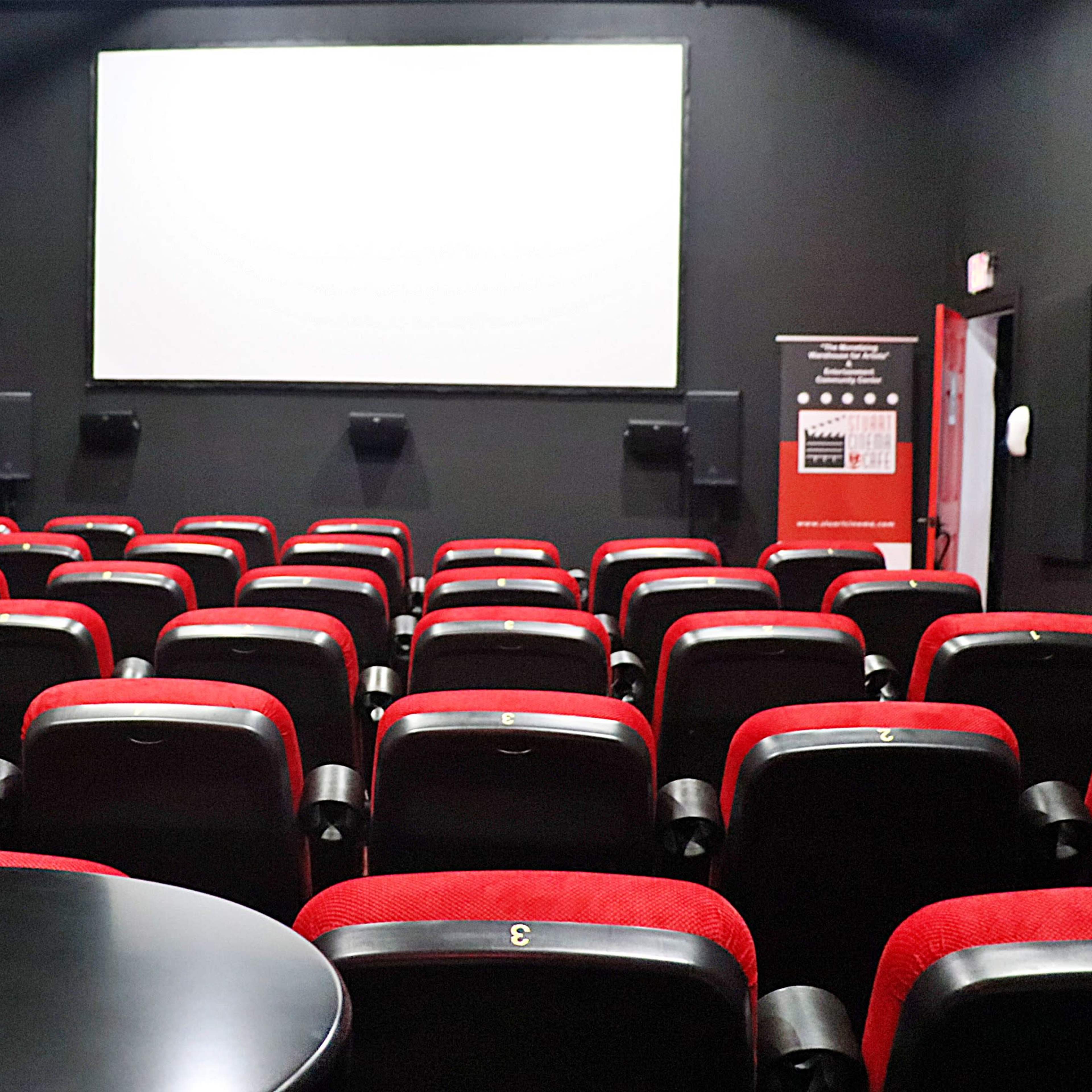 A nearly empty cinema room with rows of red and black seats facing a large white screen.
