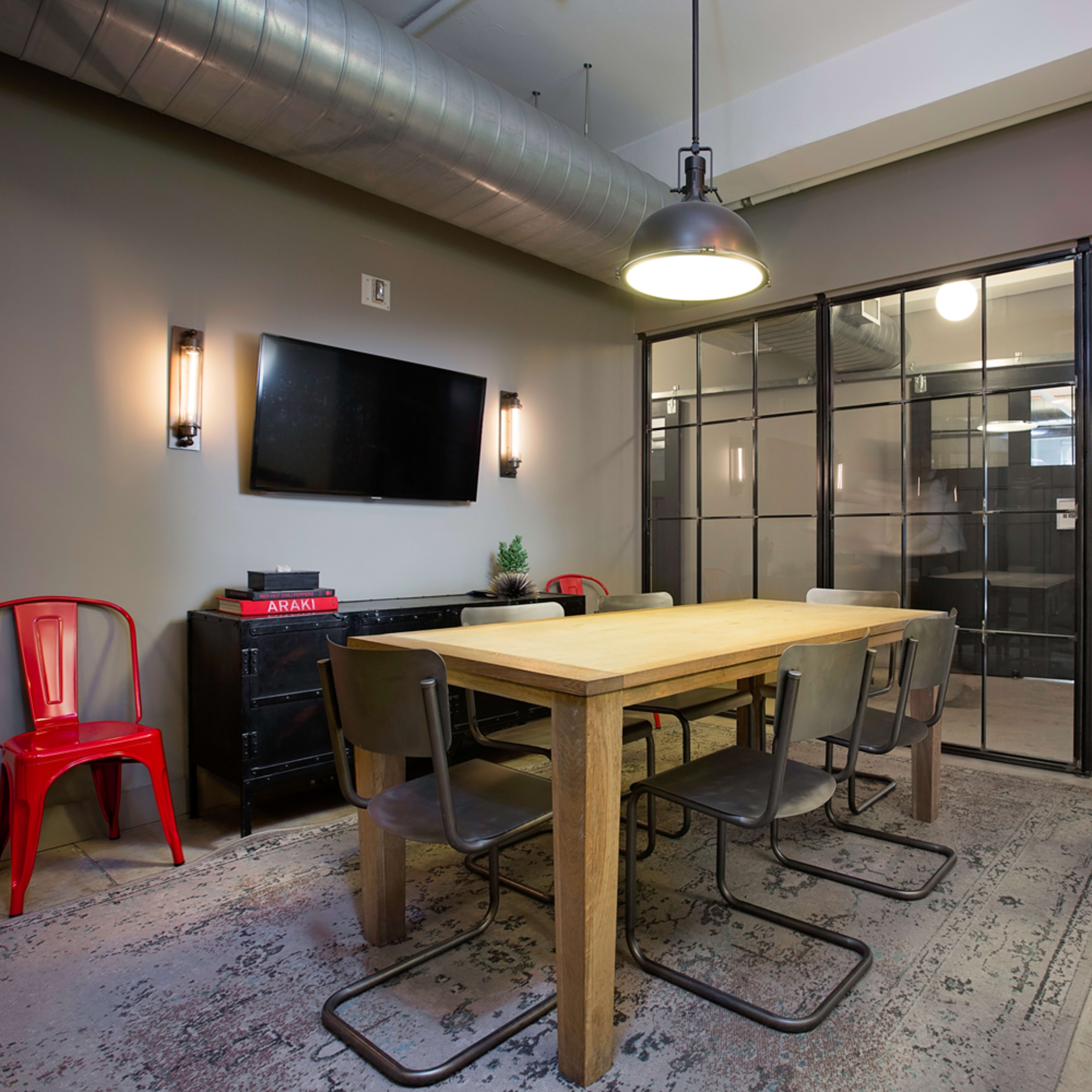 A conference room features a wooden table surrounded by metal chairs, with a large glass partition and a wall-mounted TV.