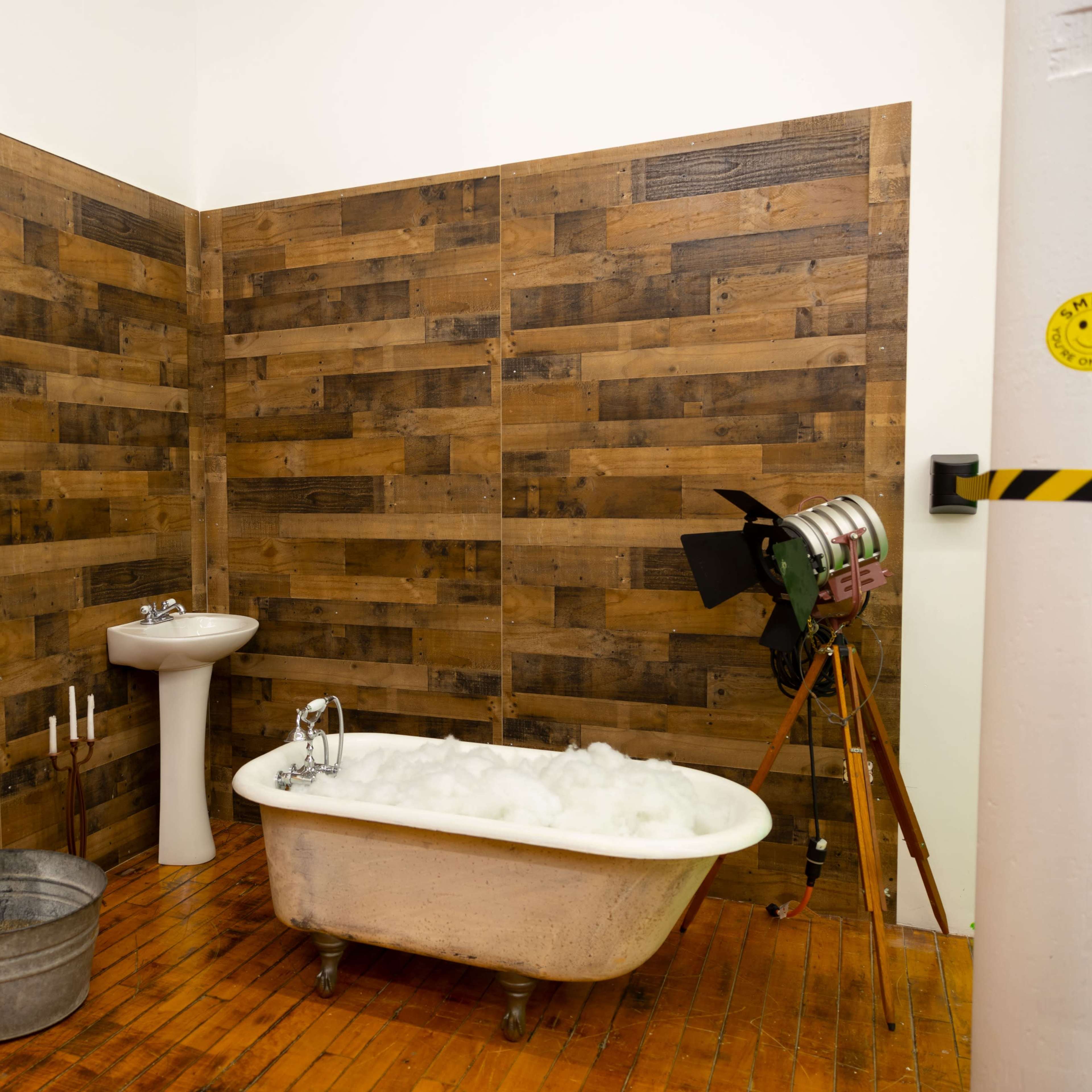 A bathroom setting with a freestanding bathtub filled with bubbles, a pedestal sink, a metal bucket, and a vintage tripod camera against a backdrop of wooden walls.