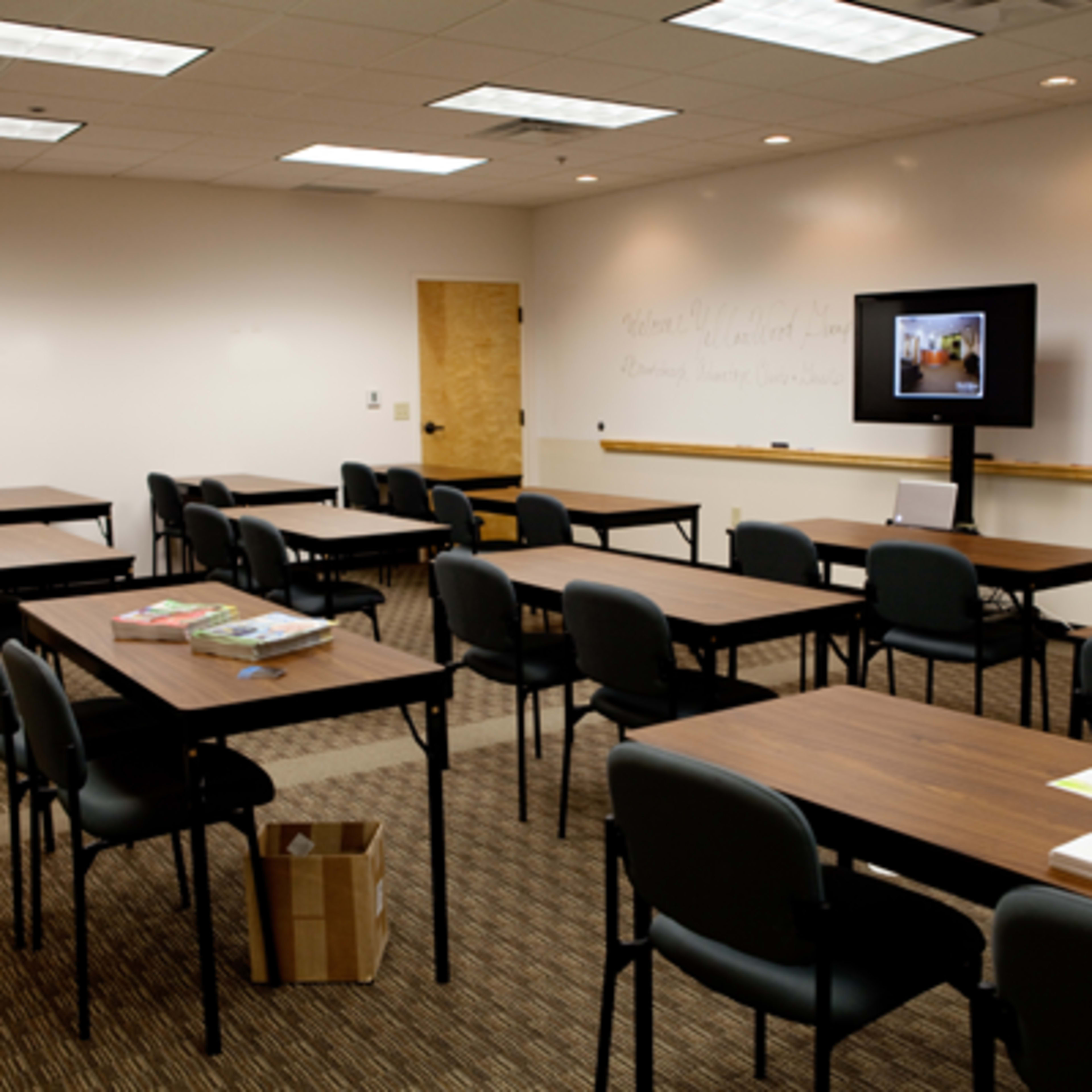 A classroom featuring multiple tables and chairs arranged in rows, with a television mounted on the wall and notes written on the whiteboard.