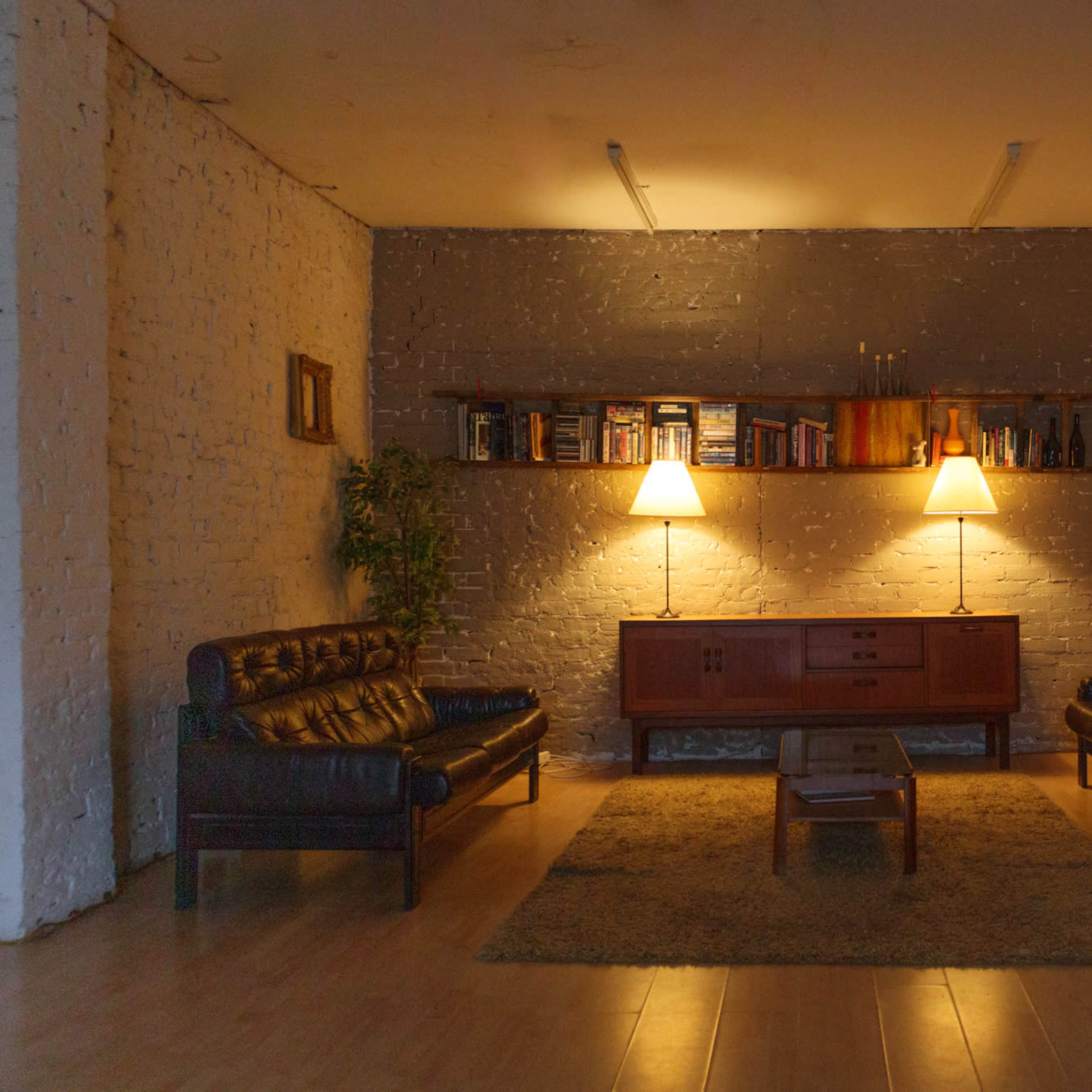 A cozy living room features two leather sofas and two armchairs arranged around a central coffee table, illuminated by two lamps against a textured gray wall.