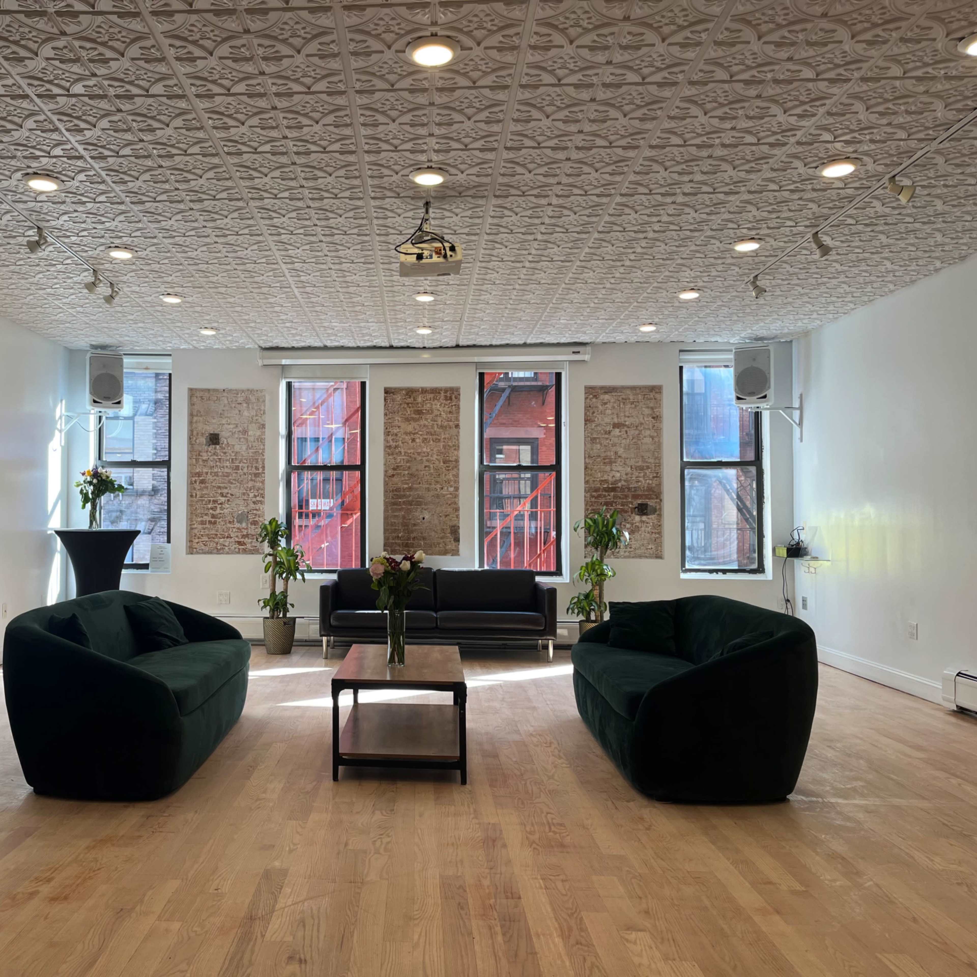 A spacious, well-lit room featuring two green upholstered armchairs, a coffee table, a black sofa, and large windows with planters.