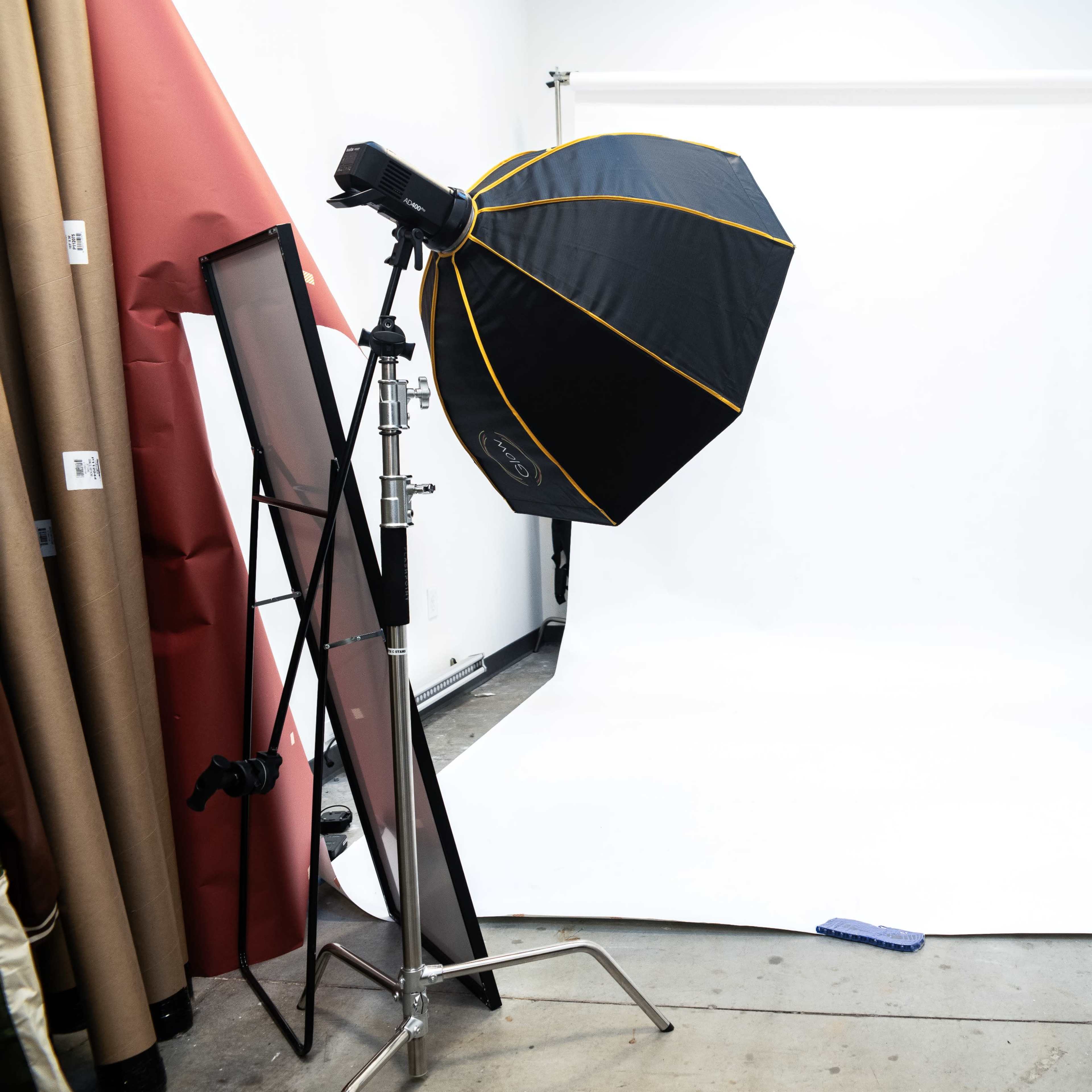 A photography studio setup with a large softbox light positioned beside a white backdrop and various props, including mirrors and rolls of paper, stored against the walls.