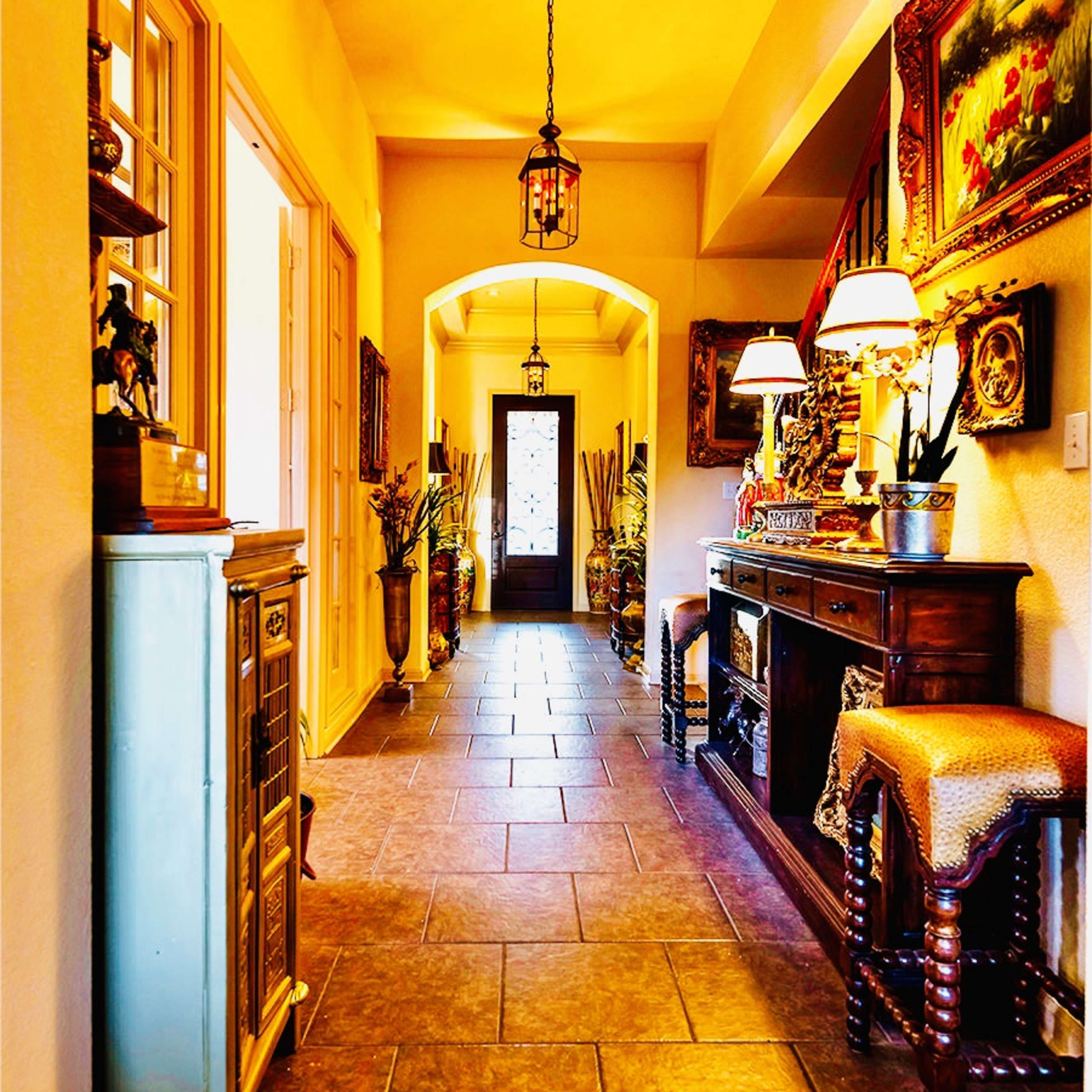 A brightly lit hallway with a tiled floor, decorated with various plants, artwork, and furniture on each side.