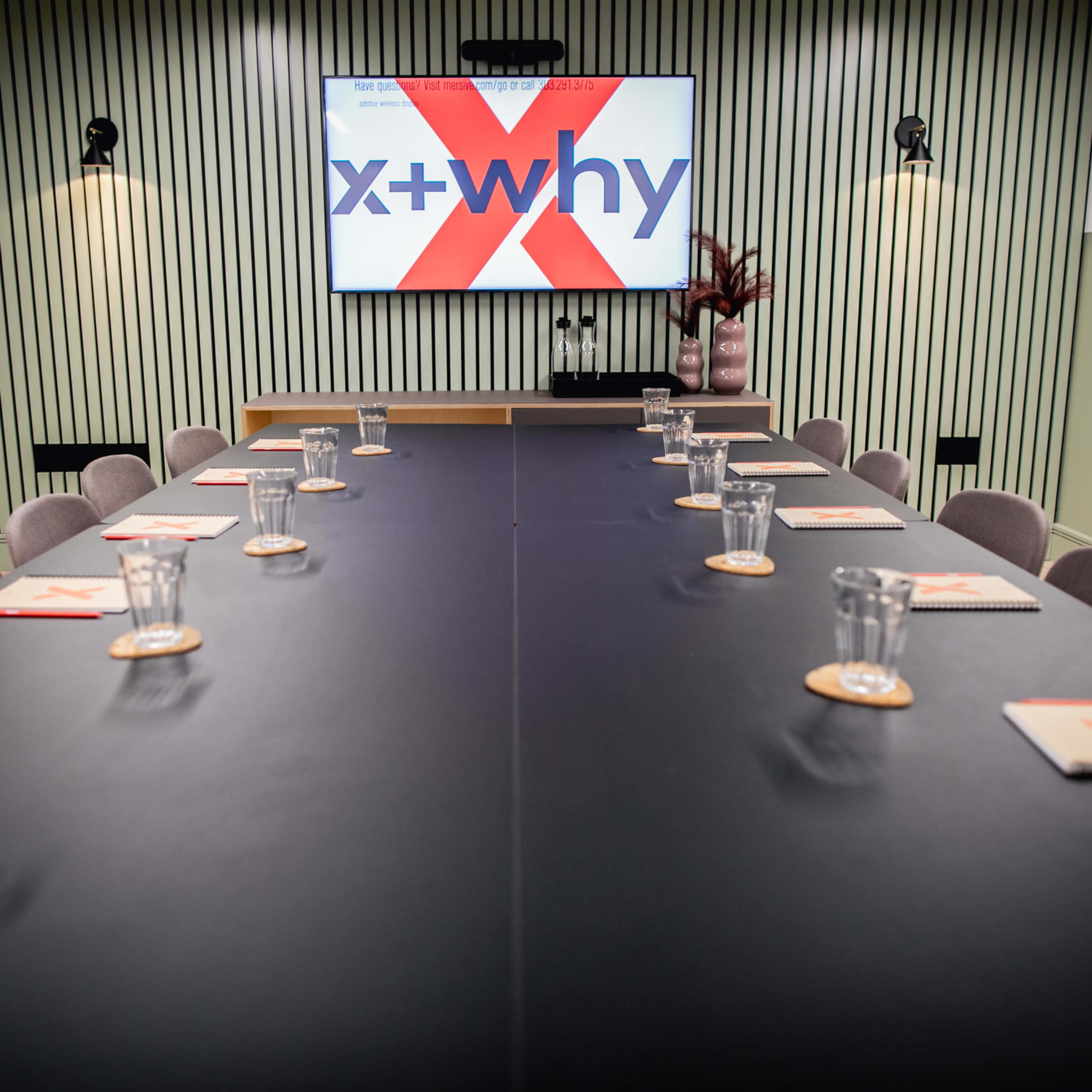 A modern conference room features a long table set with notebooks and glasses, with a large screen displaying the text "x + why" on one wall.