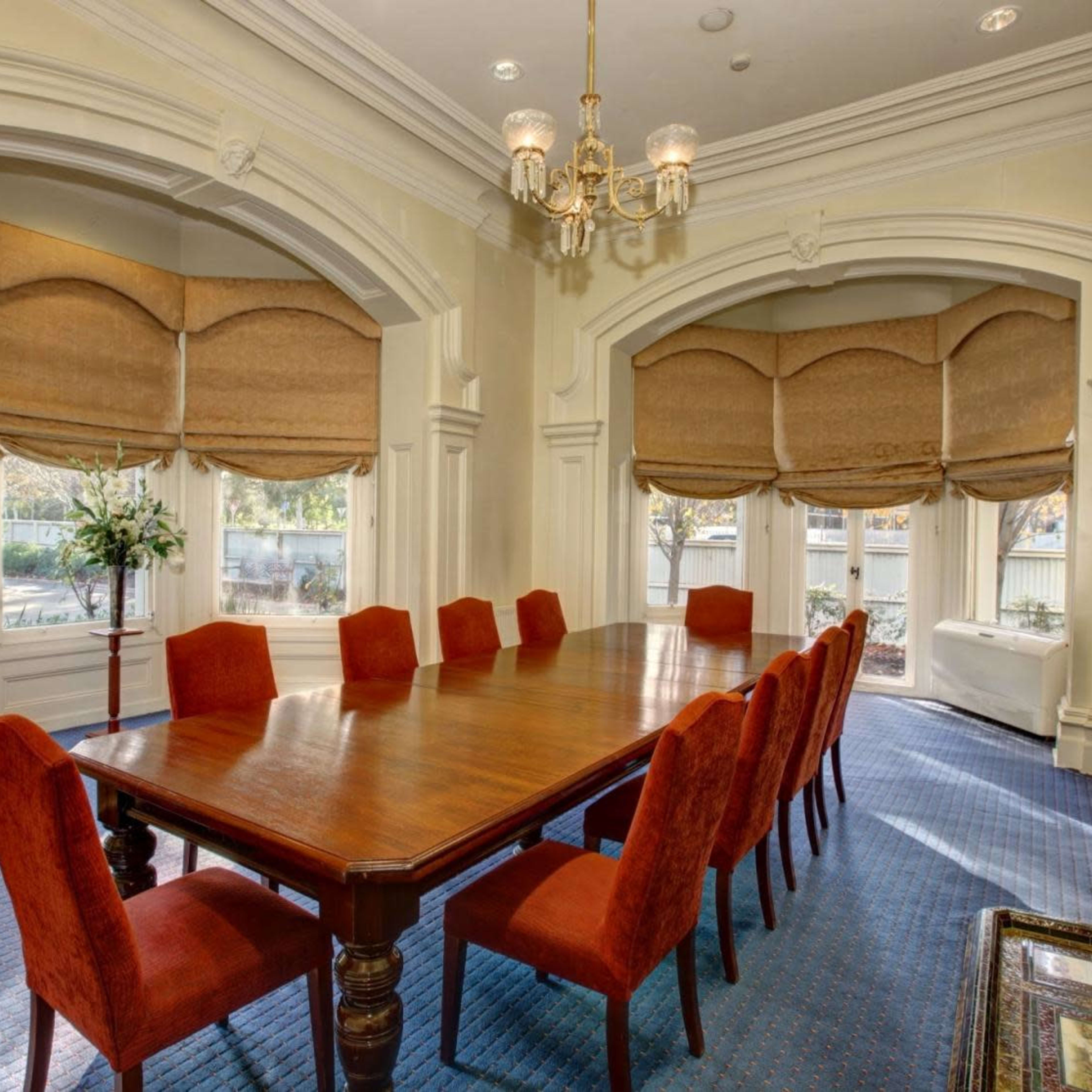 A long wooden dining table surrounded by red upholstered chairs is positioned in a well-lit room with large windows and decorative window treatments.