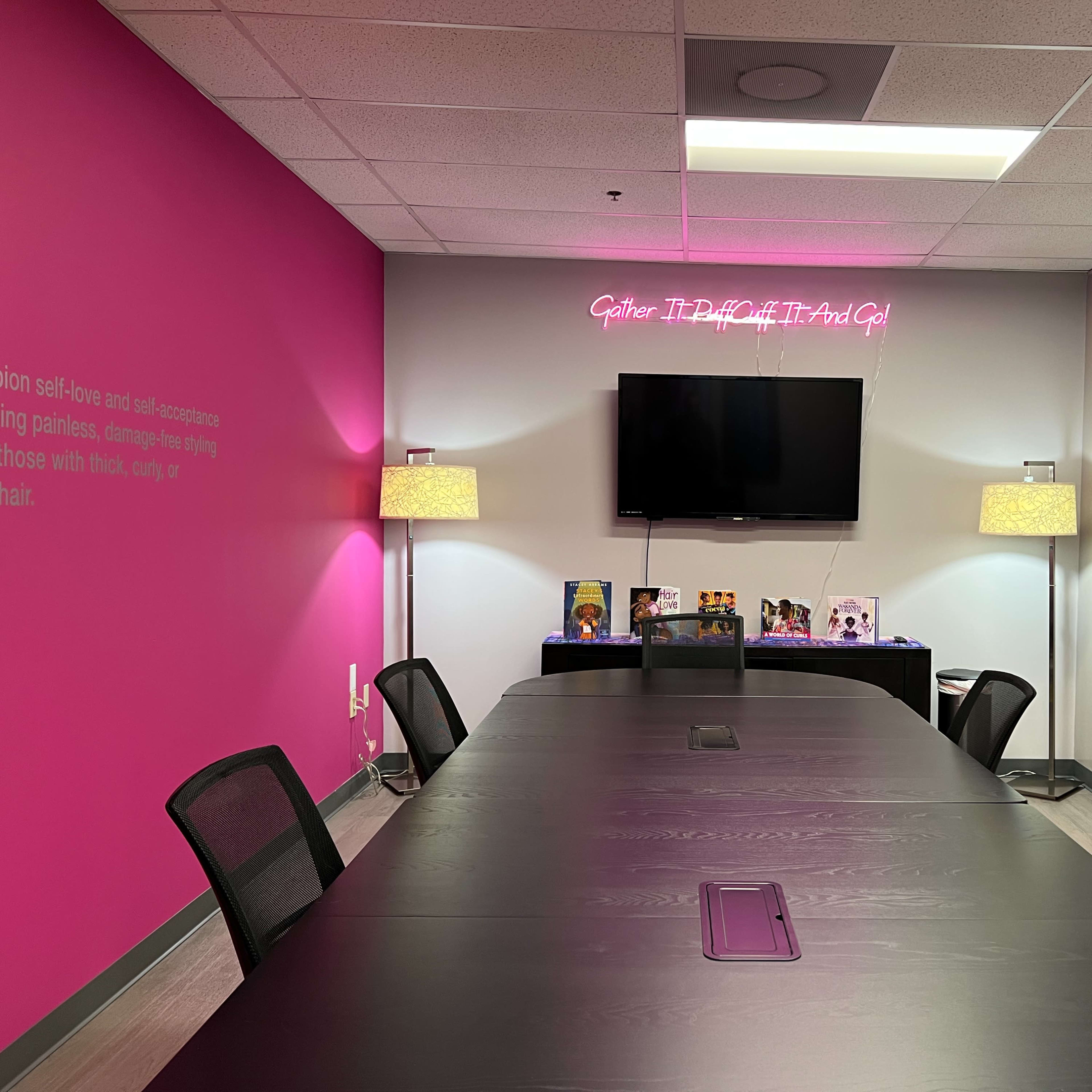 A conference room features a long black table, two standing lamps, a wall-mounted TV, and a pink accent wall with decorative items displayed on a table.