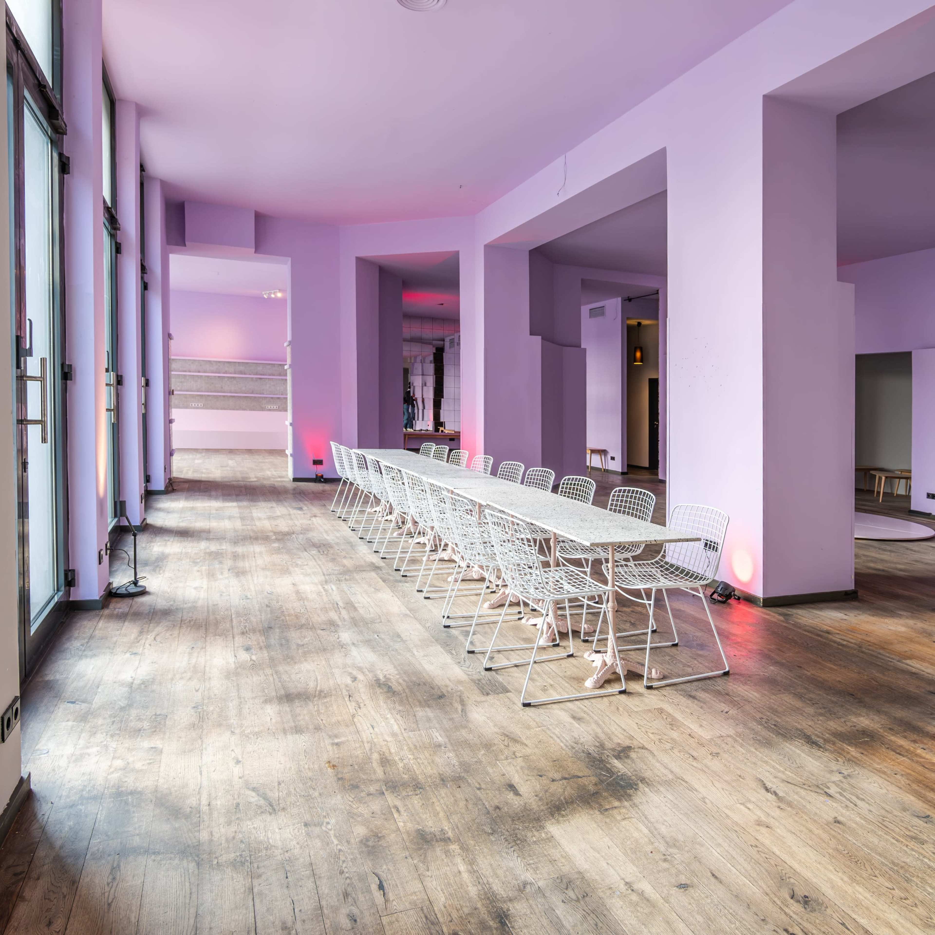 A large, empty room features a long table set with white chairs, complemented by purple walls and wooden flooring.