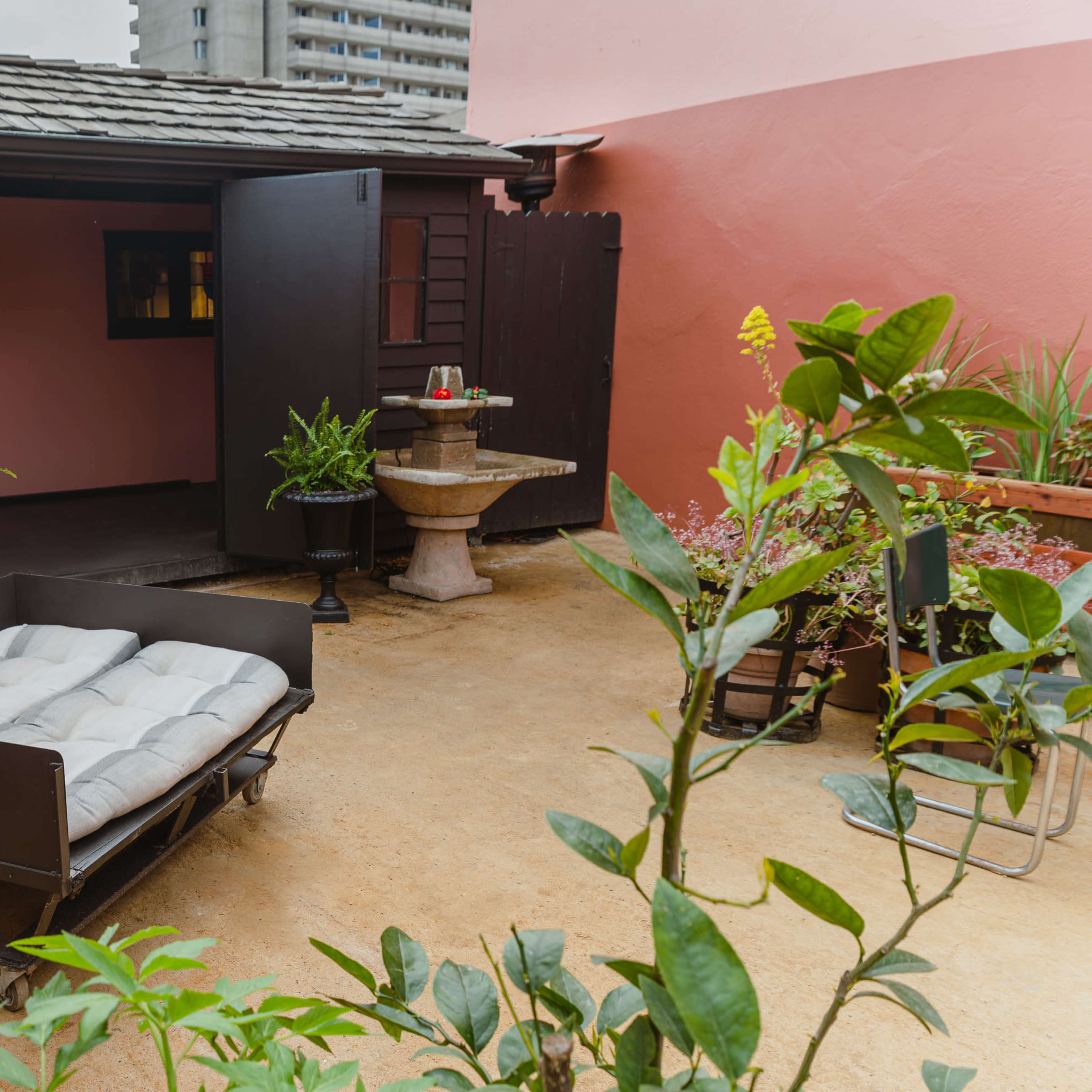 A small outdoor area features a black wooden cart with cushions, surrounded by greenery and a stone pedestal.