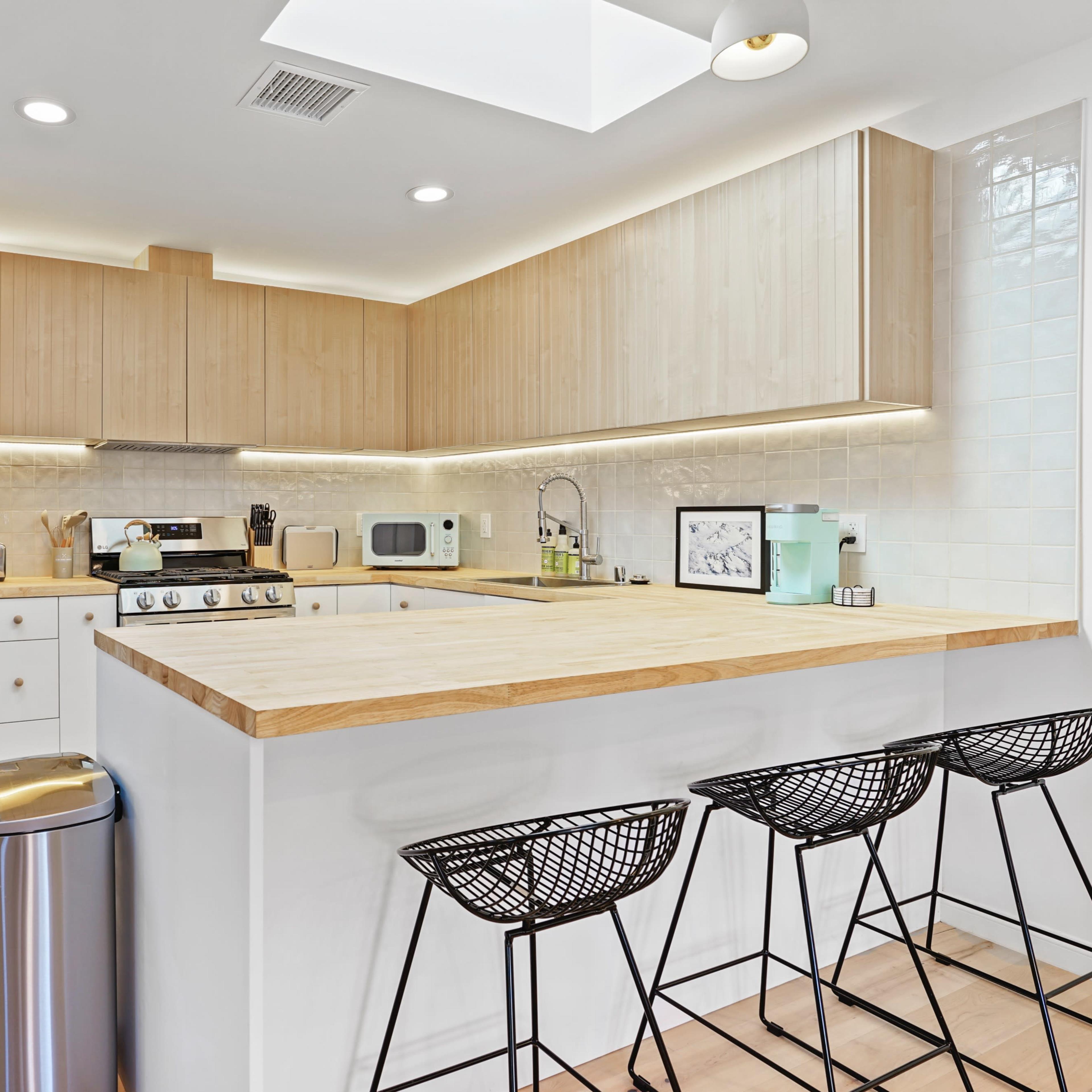 A modern kitchen featuring light wood cabinets, stainless steel appliances, a large wooden countertop, and three black wire bar stools.
