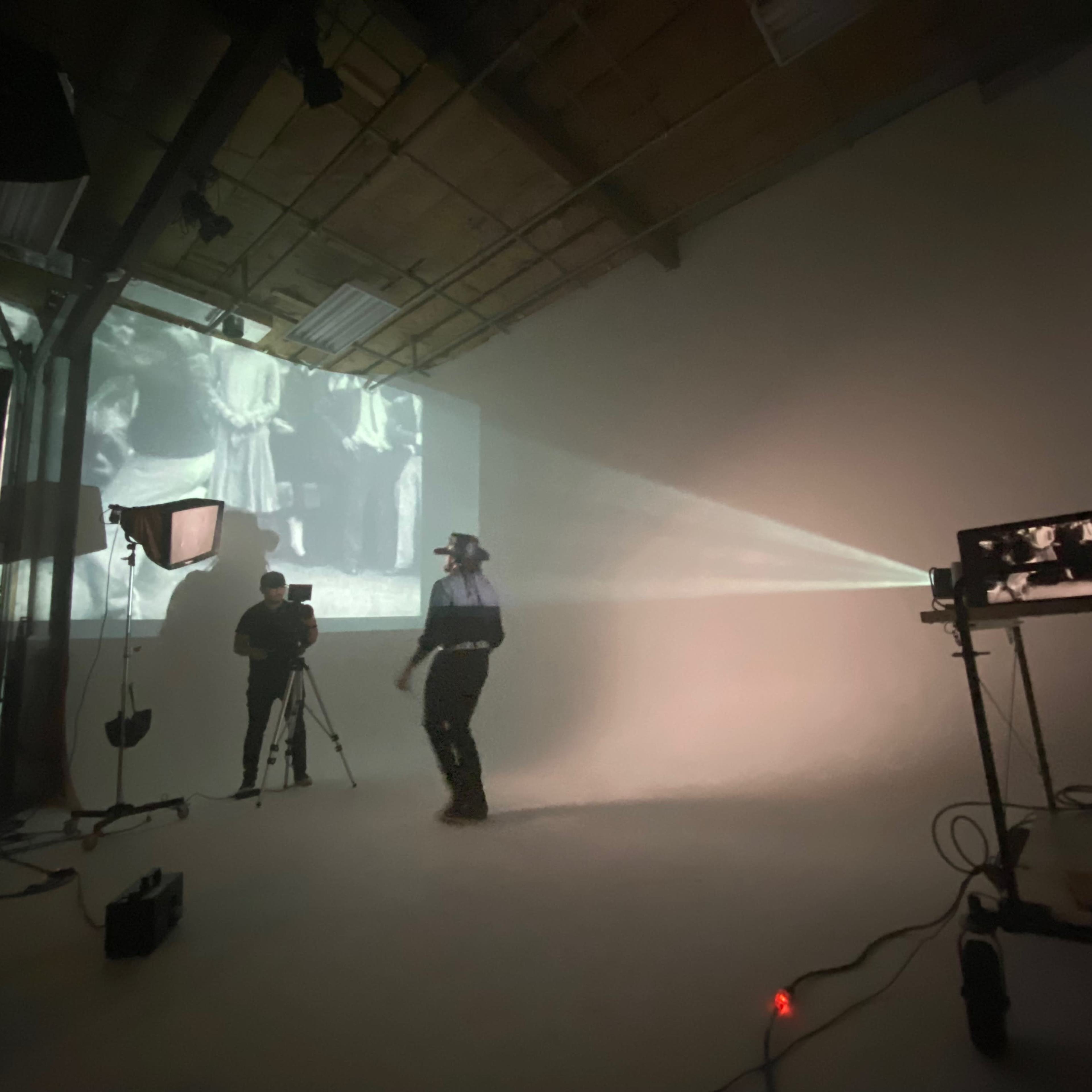A person operates a camera while another walks in front of a projected black-and-white film in a studio space.