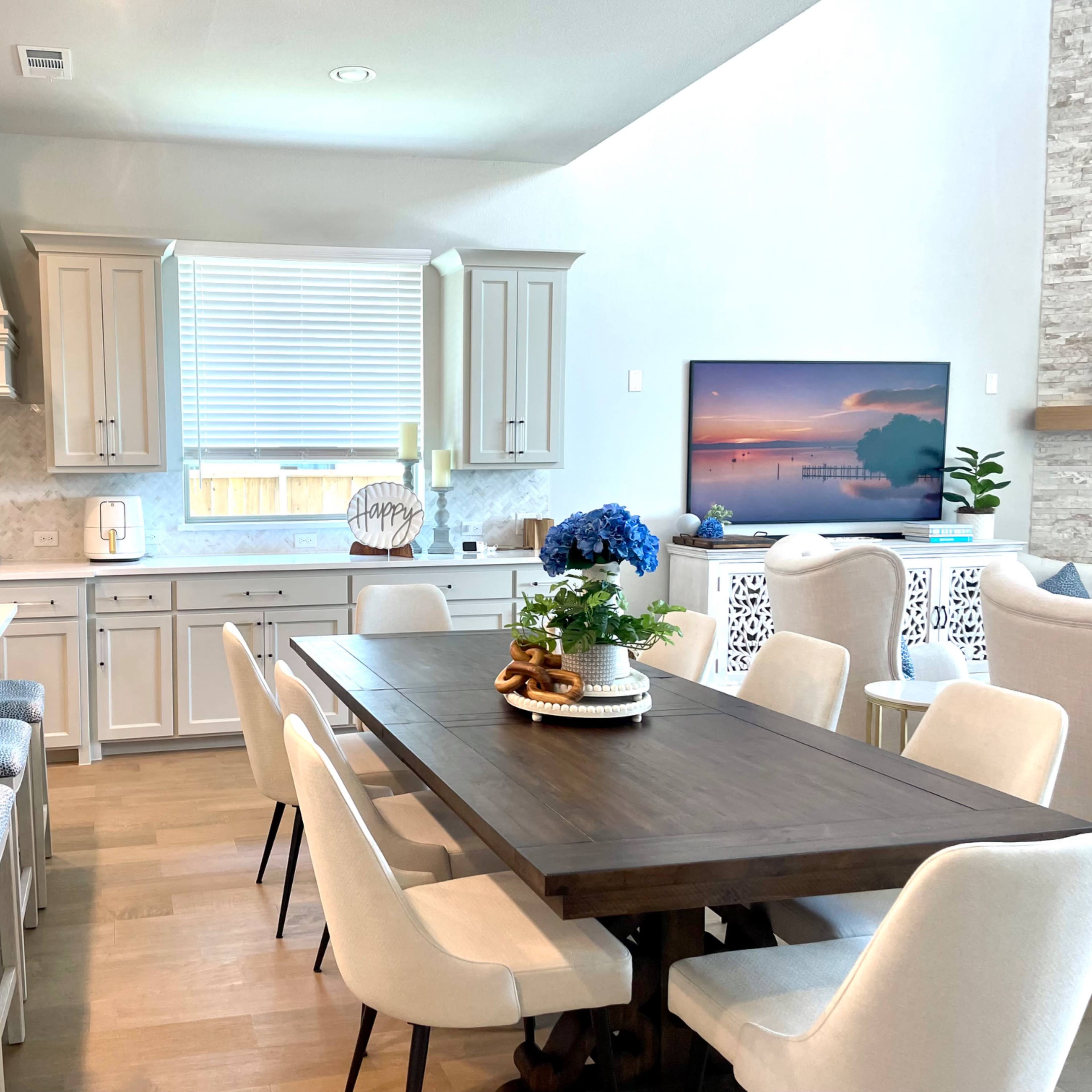 A modern kitchen and dining area with a large wooden table, light-colored cabinetry, and a television mounted on the wall.