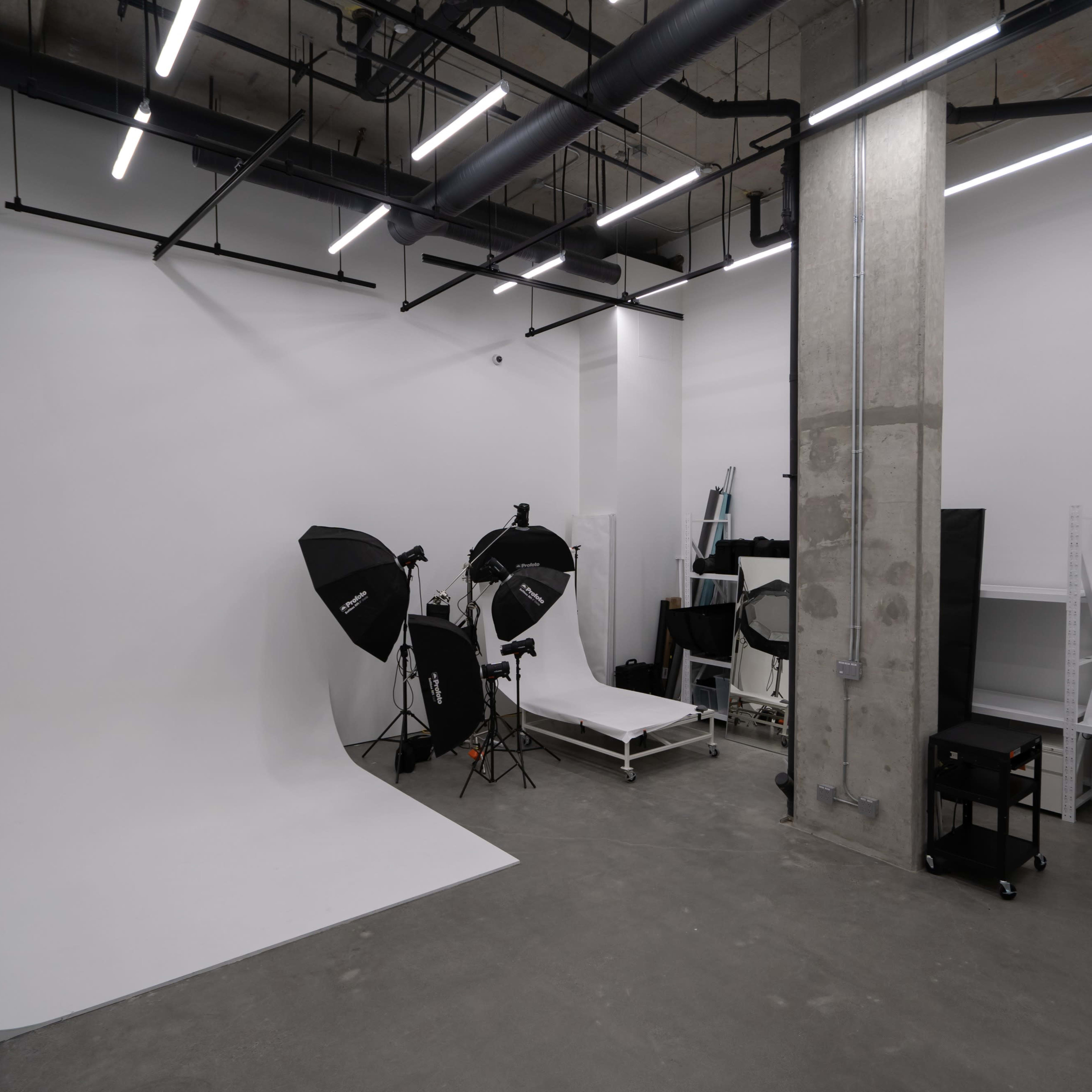 A minimalist photography studio featuring a white backdrop, lighting equipment, a rolling cart, and a workstation.