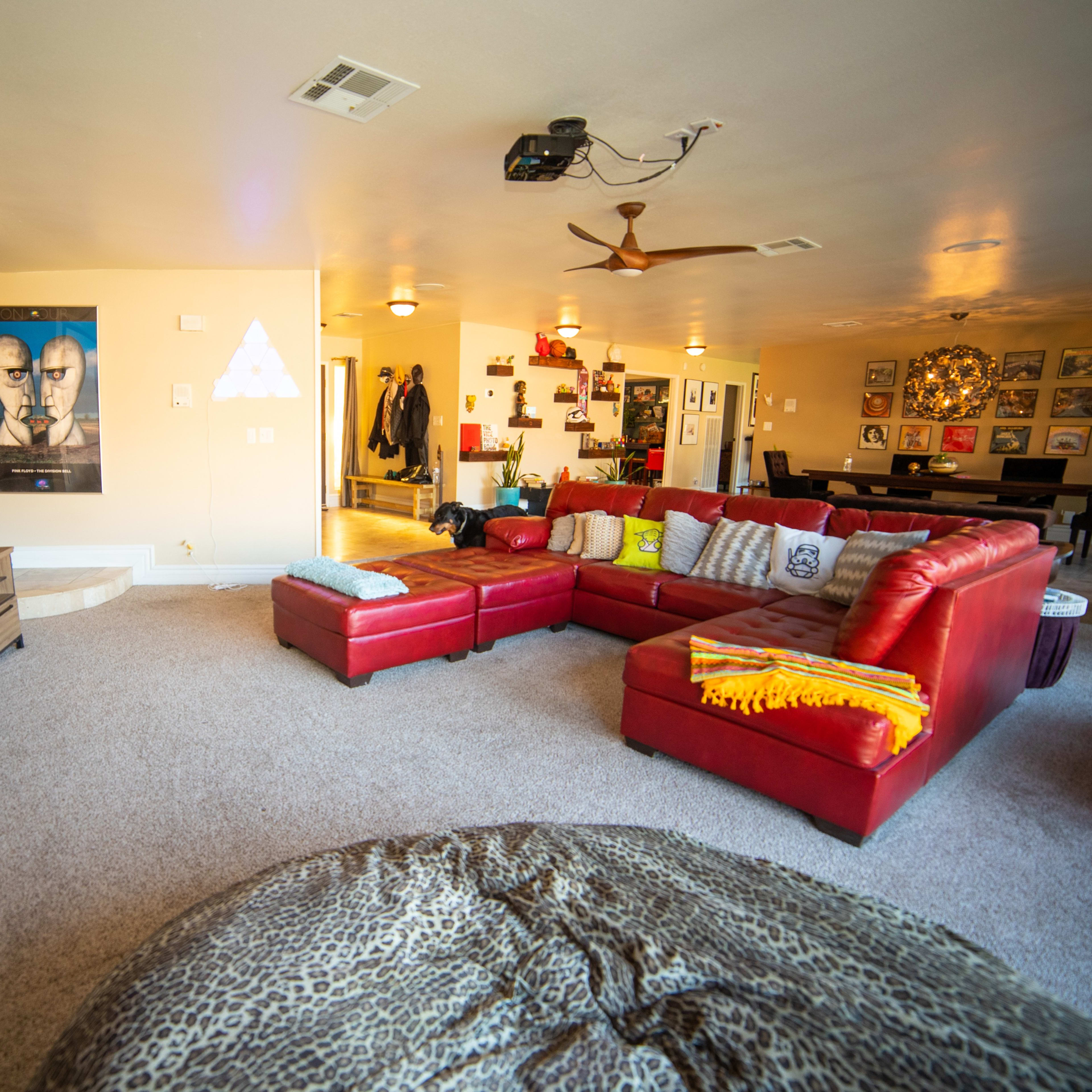 A spacious living room with a large red sectional sofa, a projector screen on the wall, and various decorations, including a poster and a round ottoman.