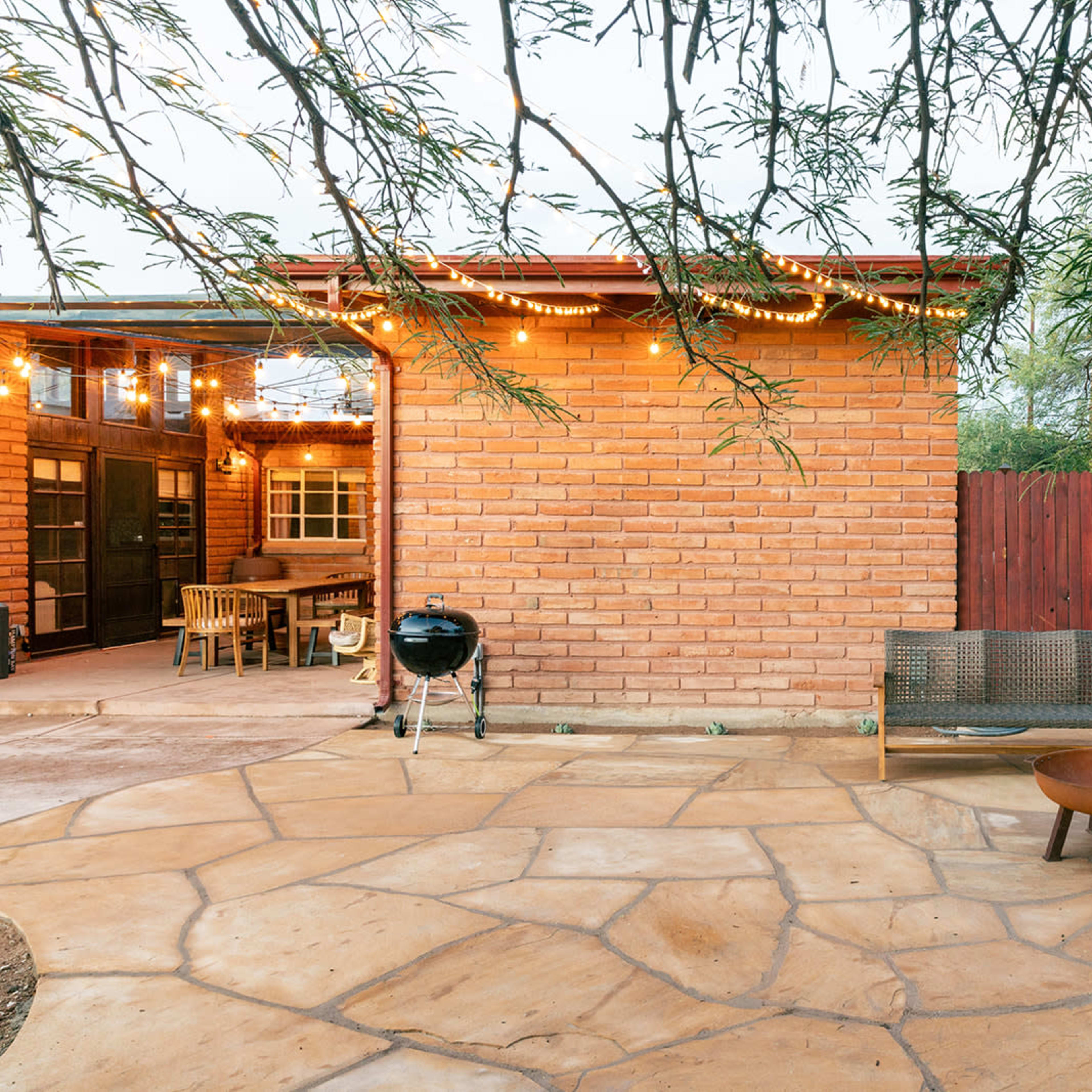 A backyard patio with stone flooring, a charcoal grill, wooden seating, and string lights suspended over a brick wall.