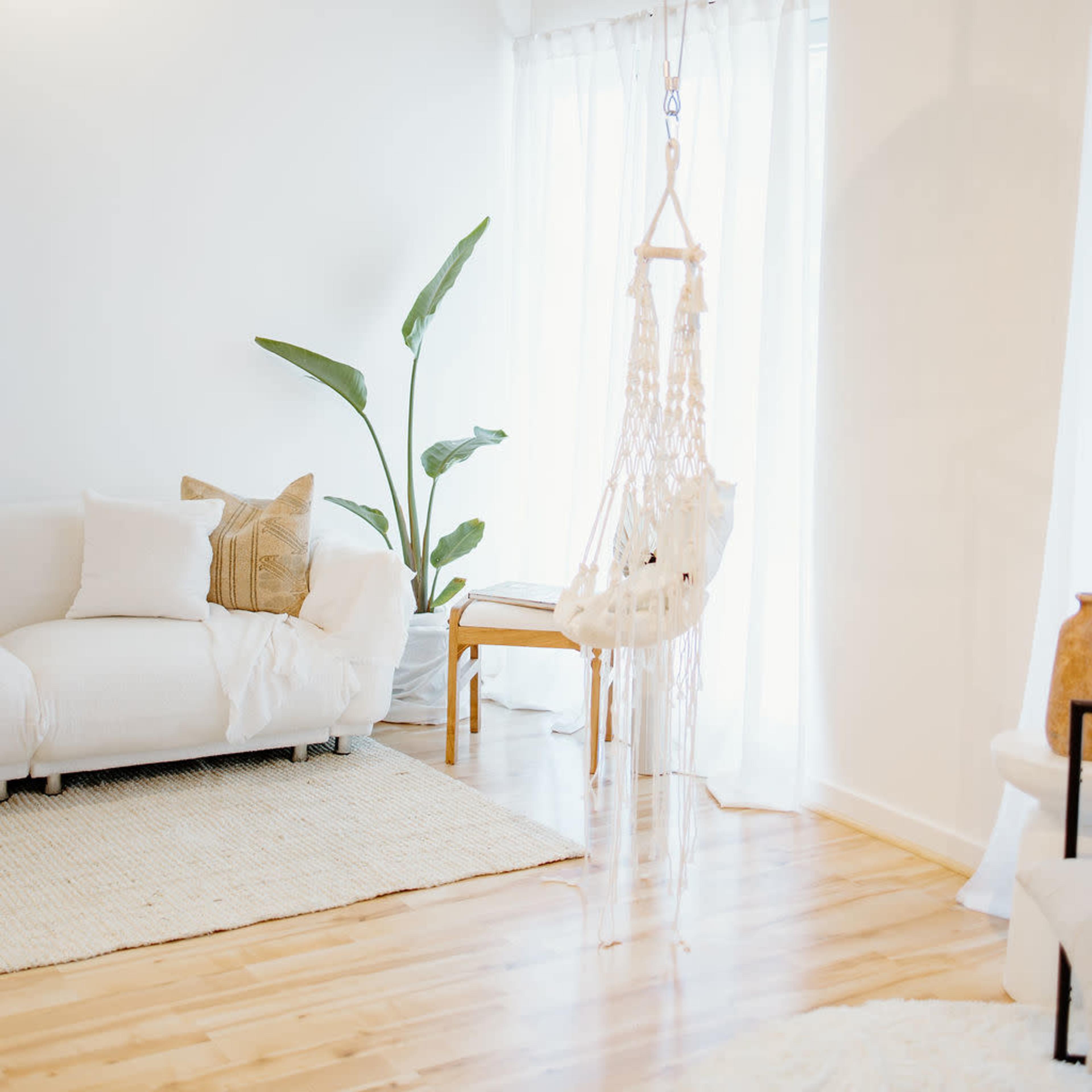 A bright, airy living space featuring a white sofa, a macramé hanging chair, and a large plant near a large window.