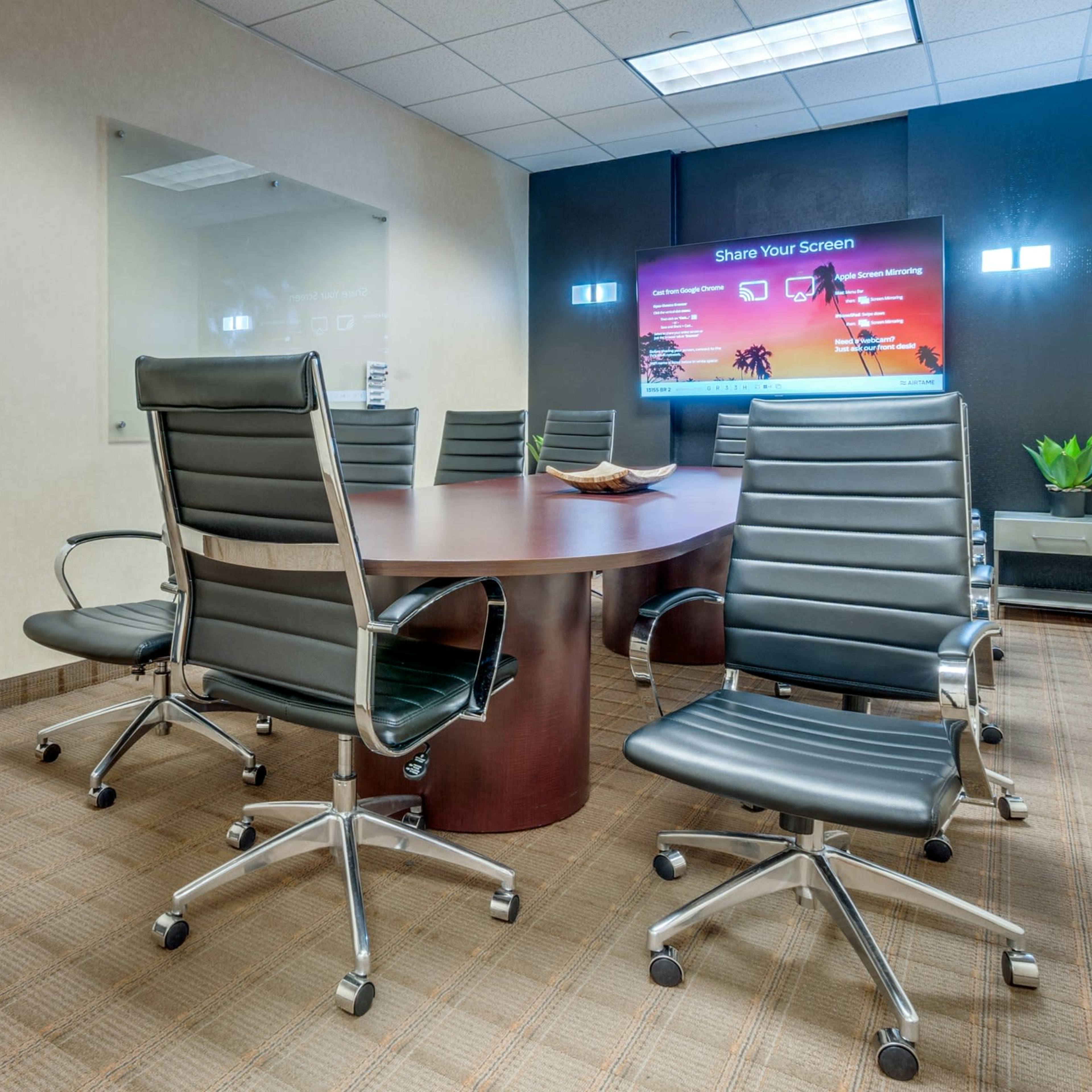 A modern conference room features a round table surrounded by ergonomic chairs and a large screen displaying a "Share Your Screen" message.