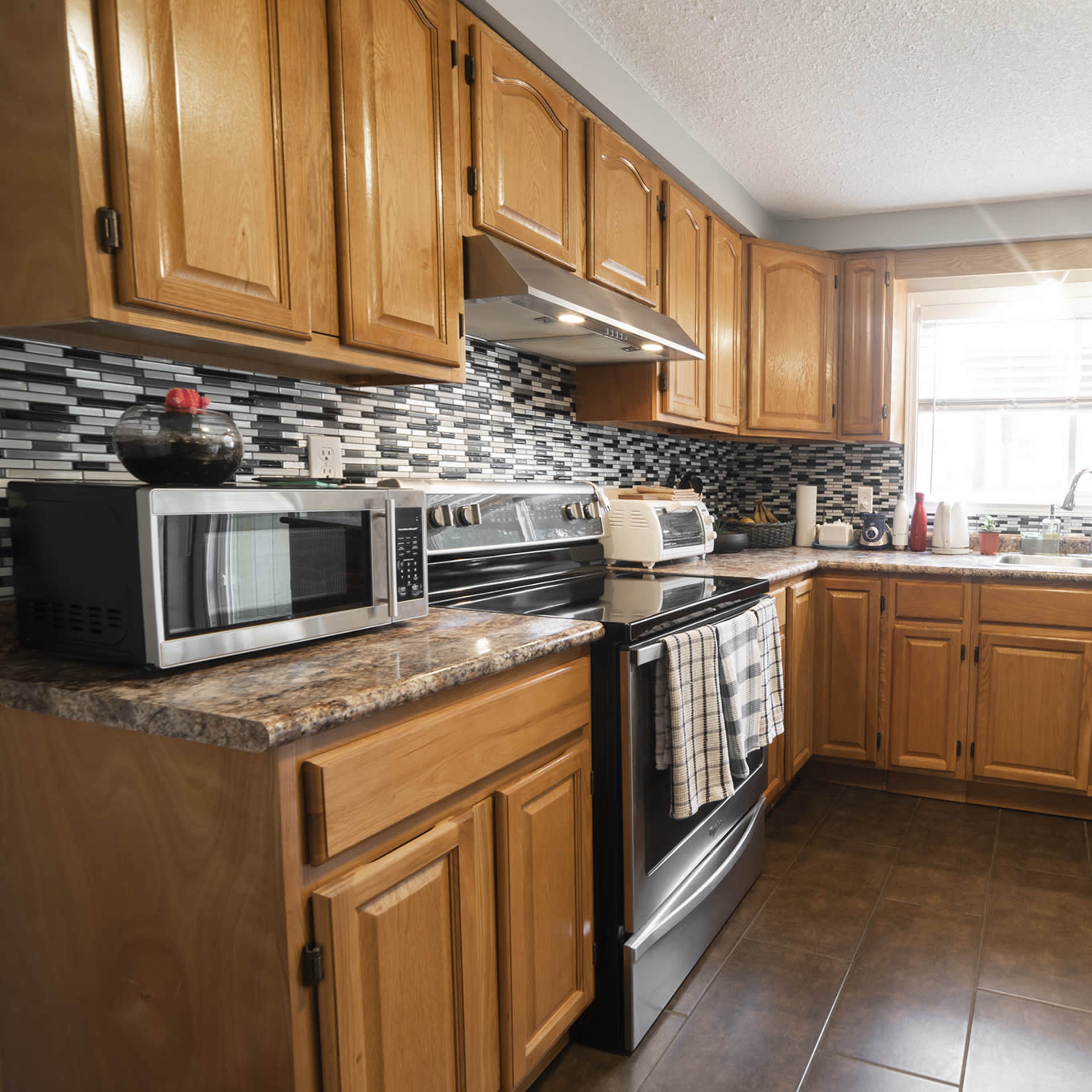 A modern kitchen with wooden cabinets, a stainless steel stove, and a microwave, featuring a tile backsplash and a large window above the sink.