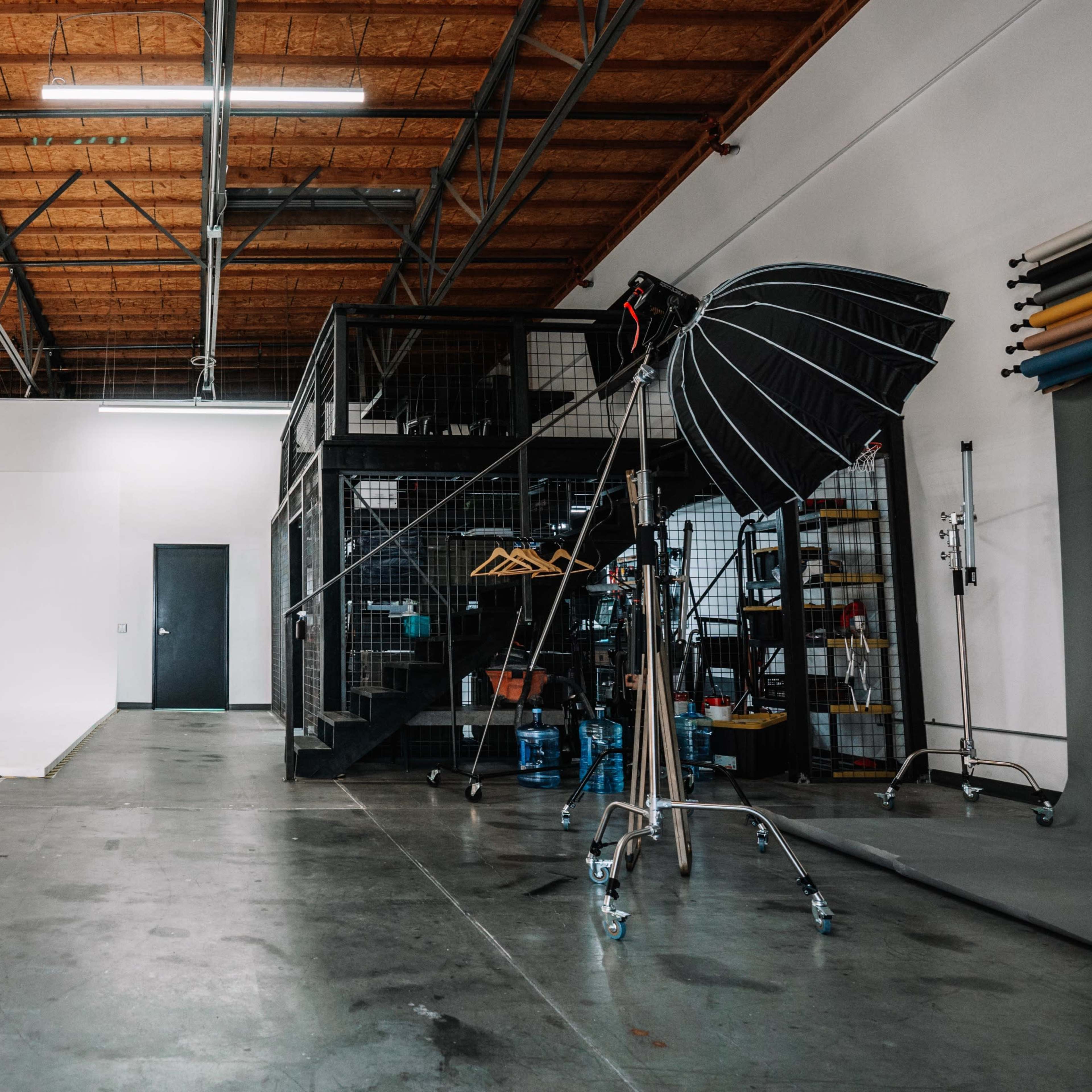 A large, empty studio space with a white backdrop, a metal staircase leading to an upper storage area, and photography equipment arranged on one side.