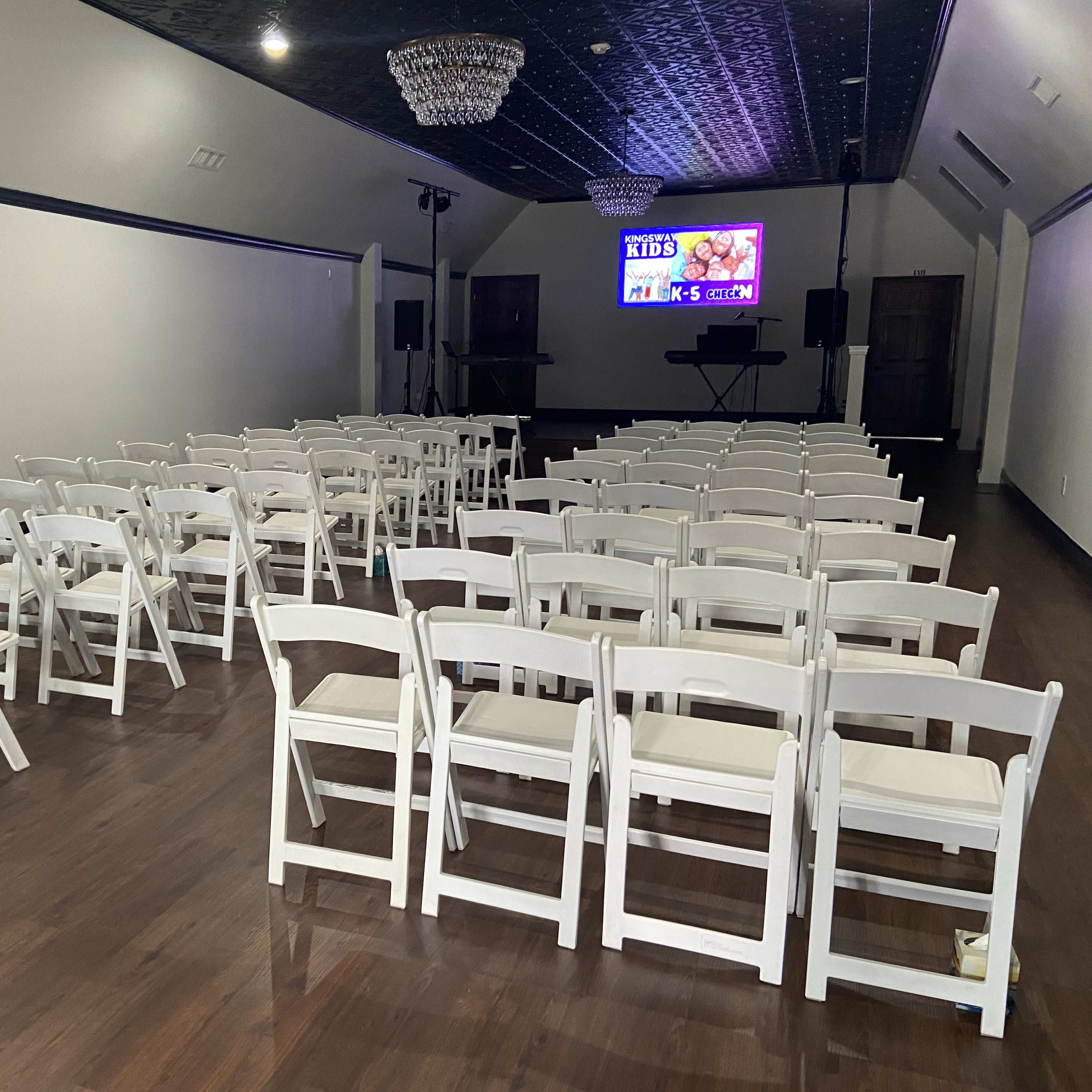 An empty event space arranged with rows of white folding chairs facing a large screen and a small stage.