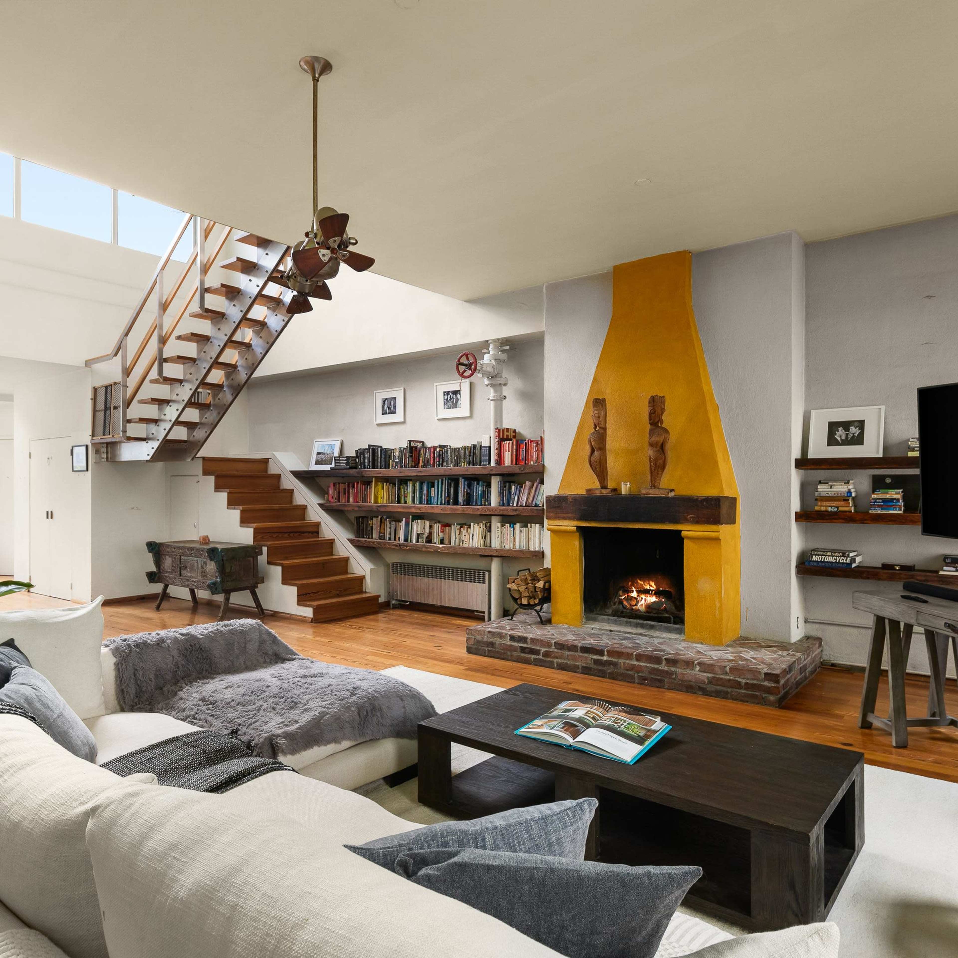 The living room features a fireplace with a yellow mantel, a large sectional sofa, a wooden coffee table, and a staircase leading to an upper level.