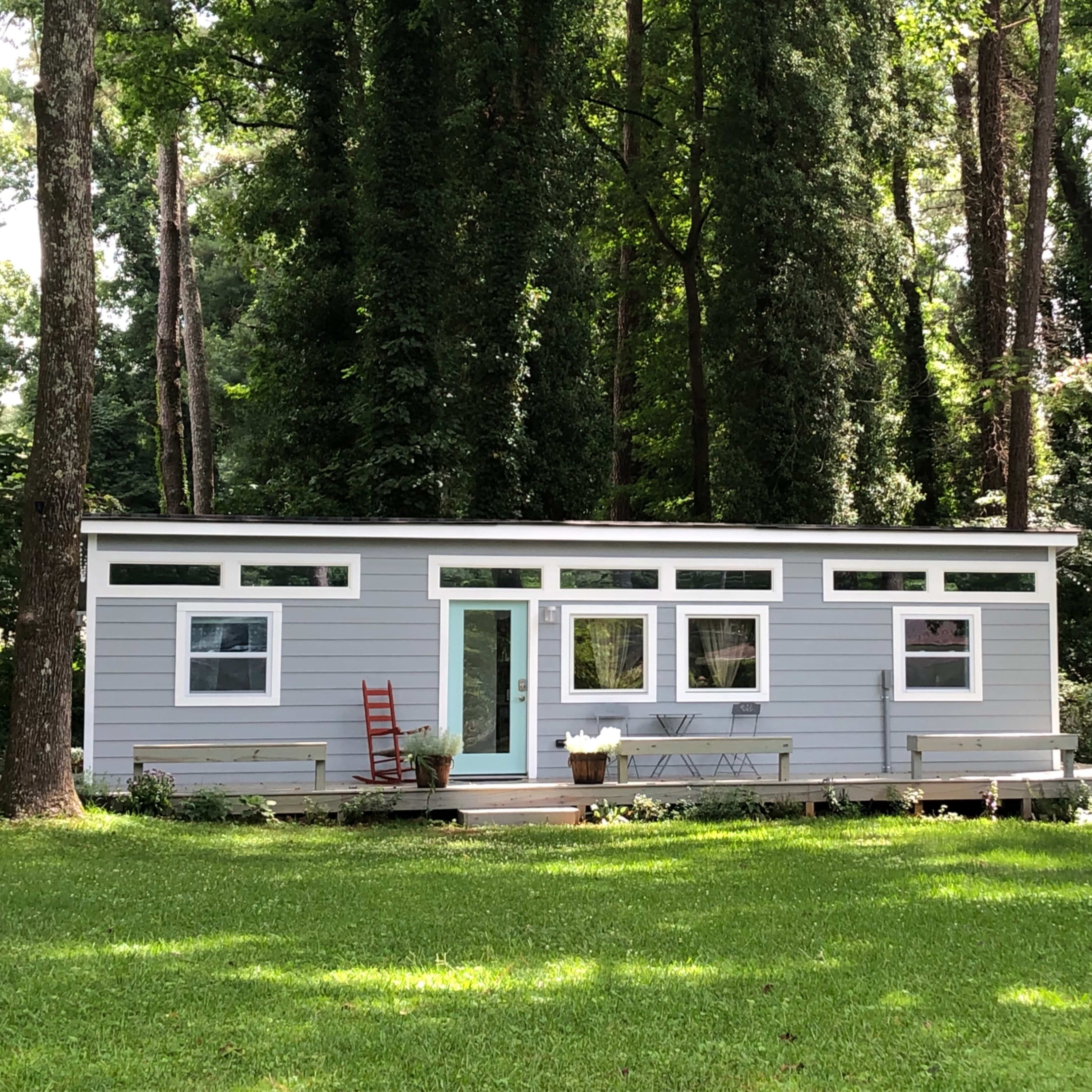 A modern, single-story cabin with large windows is set on a green lawn surrounded by tall trees.