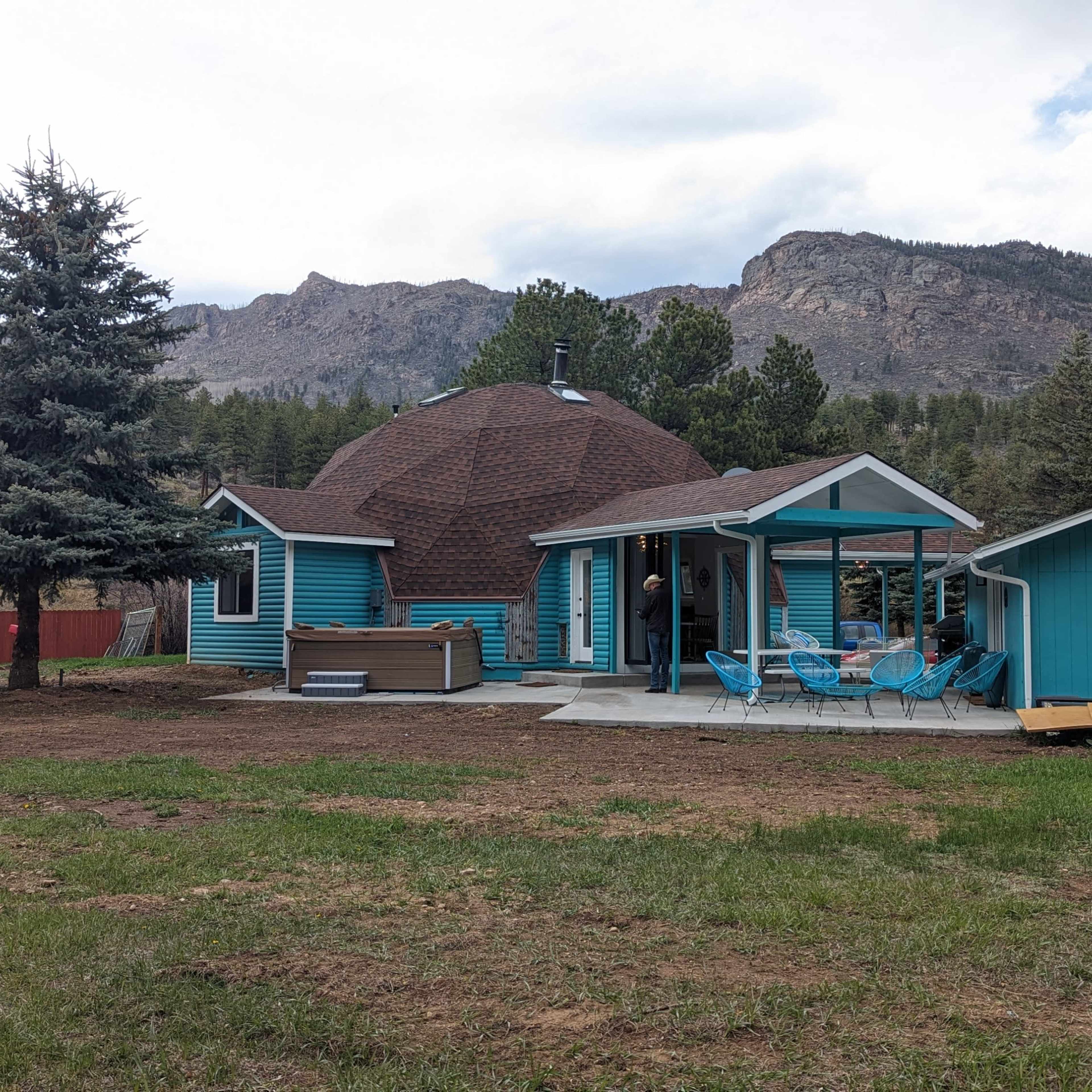 A turquoise domed house with a sloped roof and a small adjacent building is set against a backdrop of mountains and trees.