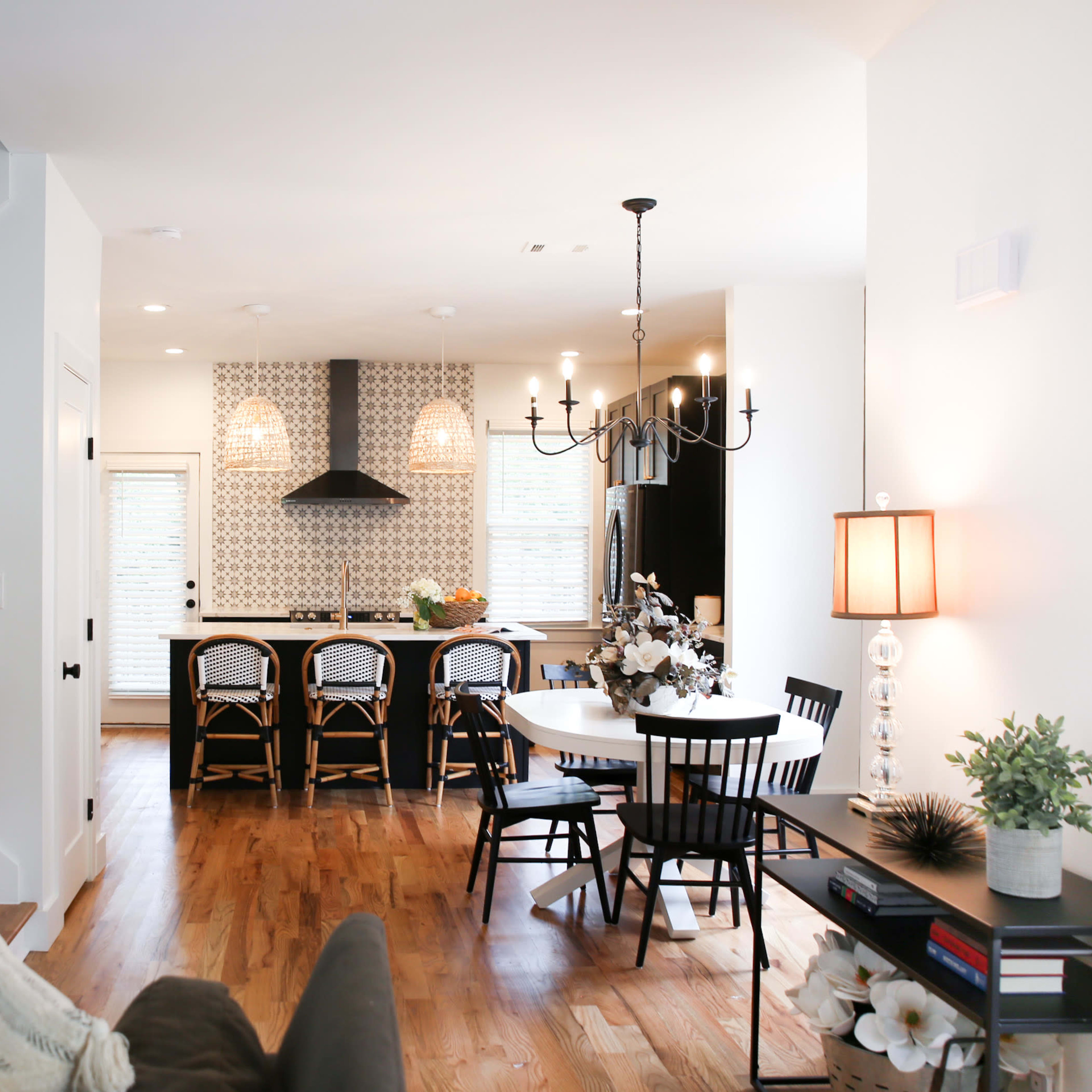 A modern open-concept living space features a staircase on the left, a dining area with a round table, and a kitchen with black cabinetry and bar seating in the background.