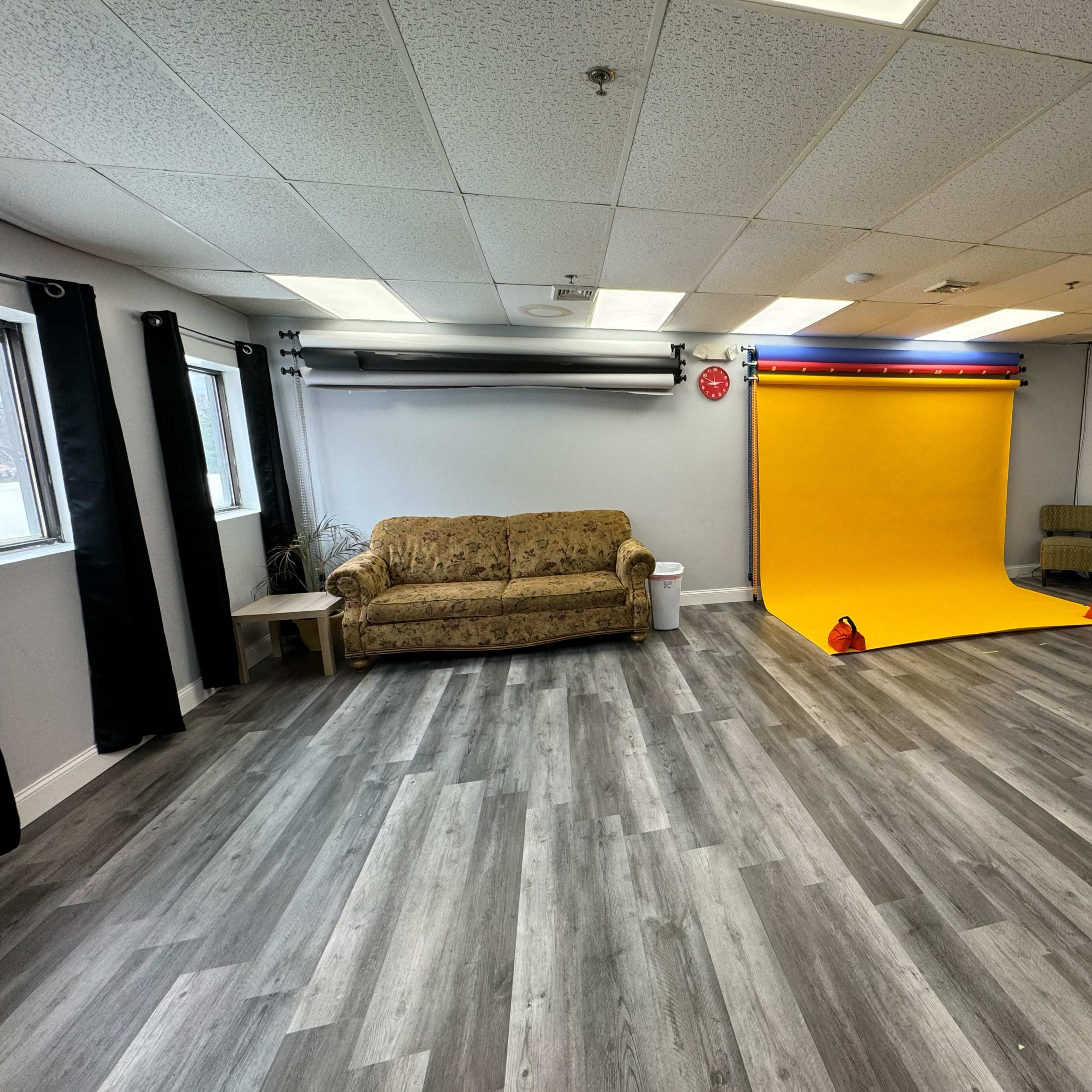 A spacious room with a yellow backdrop for photography, a patterned couch, and several windows allowing natural light.