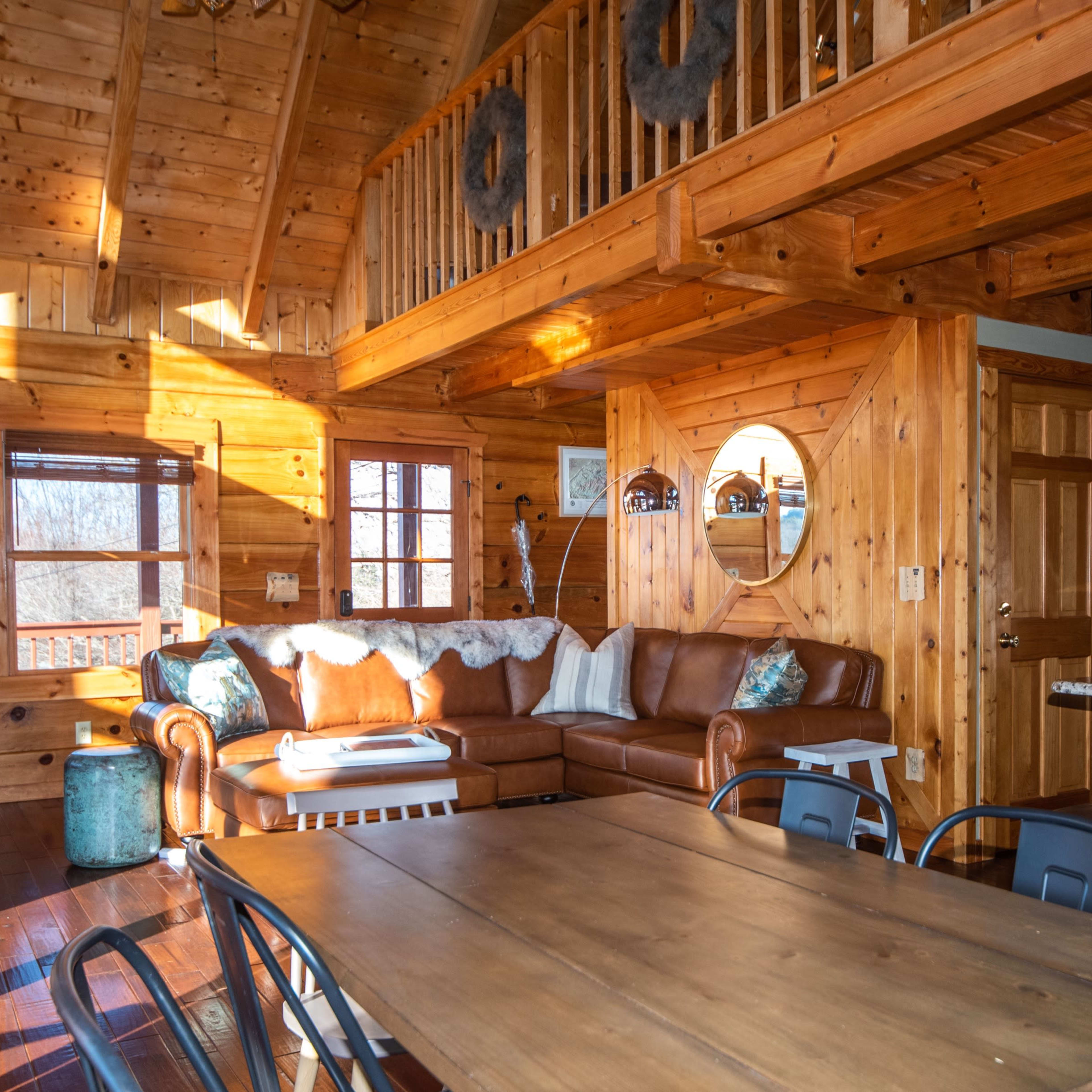 The interior of a wooden cabin features a leather sofa, a dining table, and large windows allowing sunlight to illuminate the space.