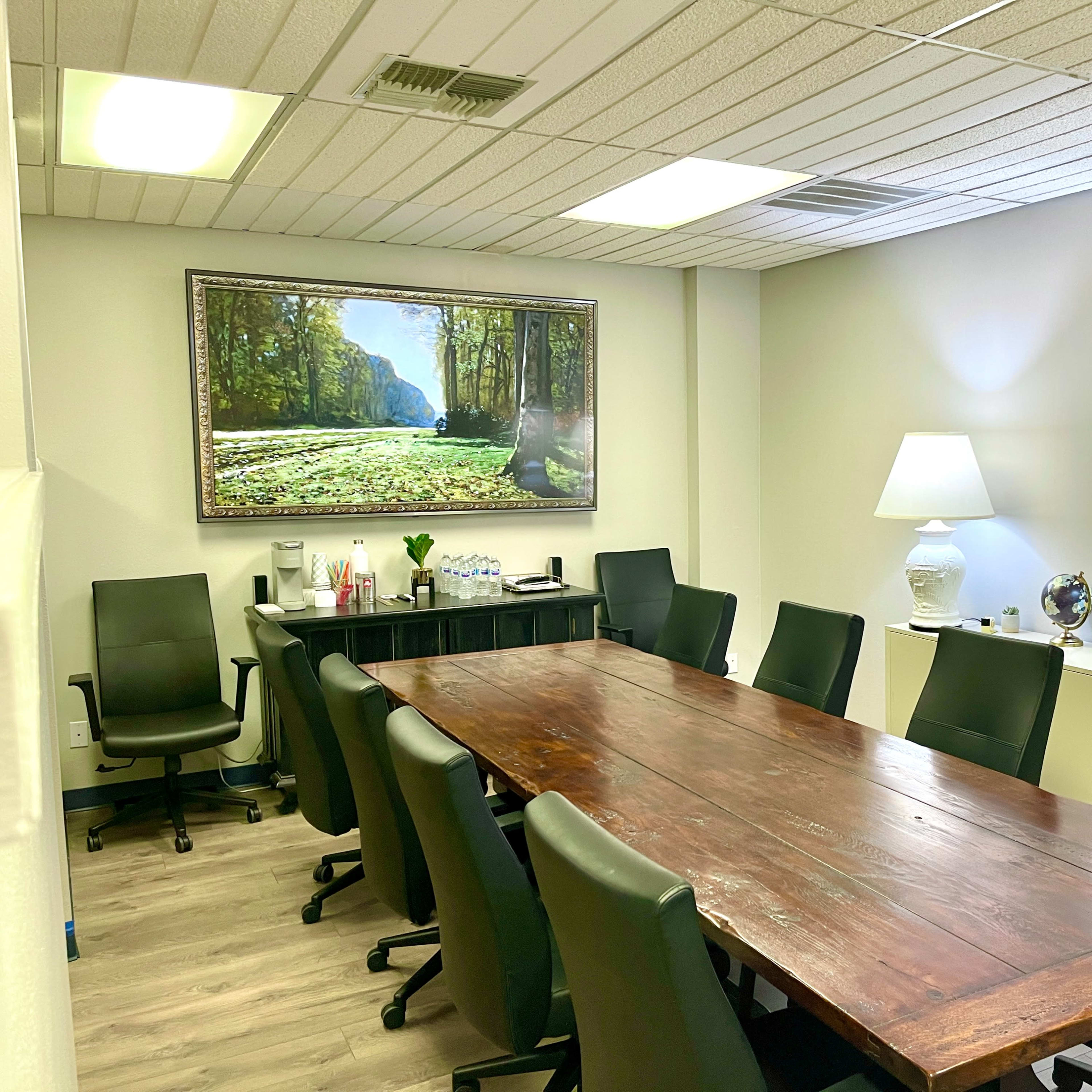 A conference room with a large wooden table, eight black chairs, two lamps, and a framed nature landscape painting on the wall.