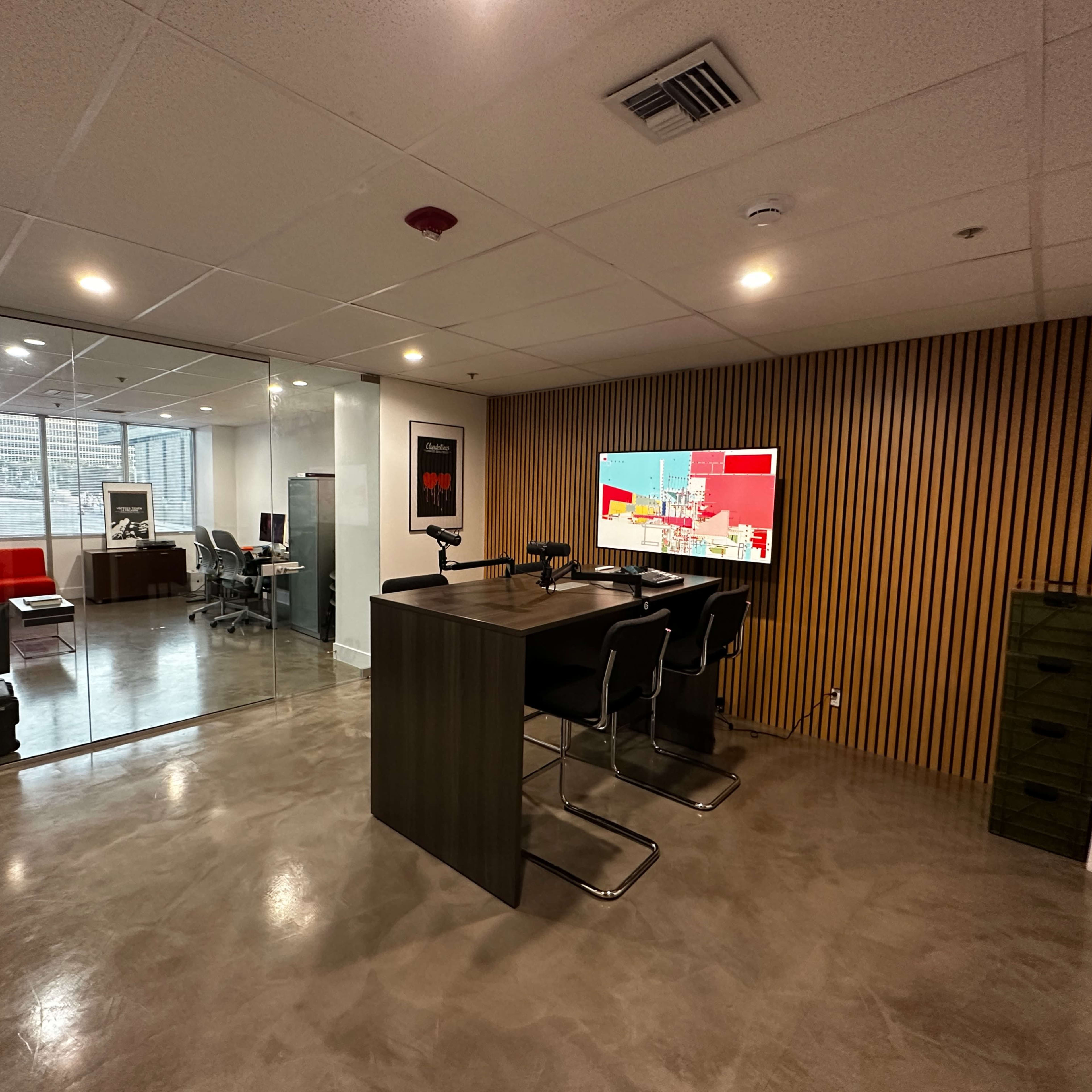 A modern office space featuring a glass-walled meeting area, a seating area with red furniture, and a workspace with a large monitor and chairs.