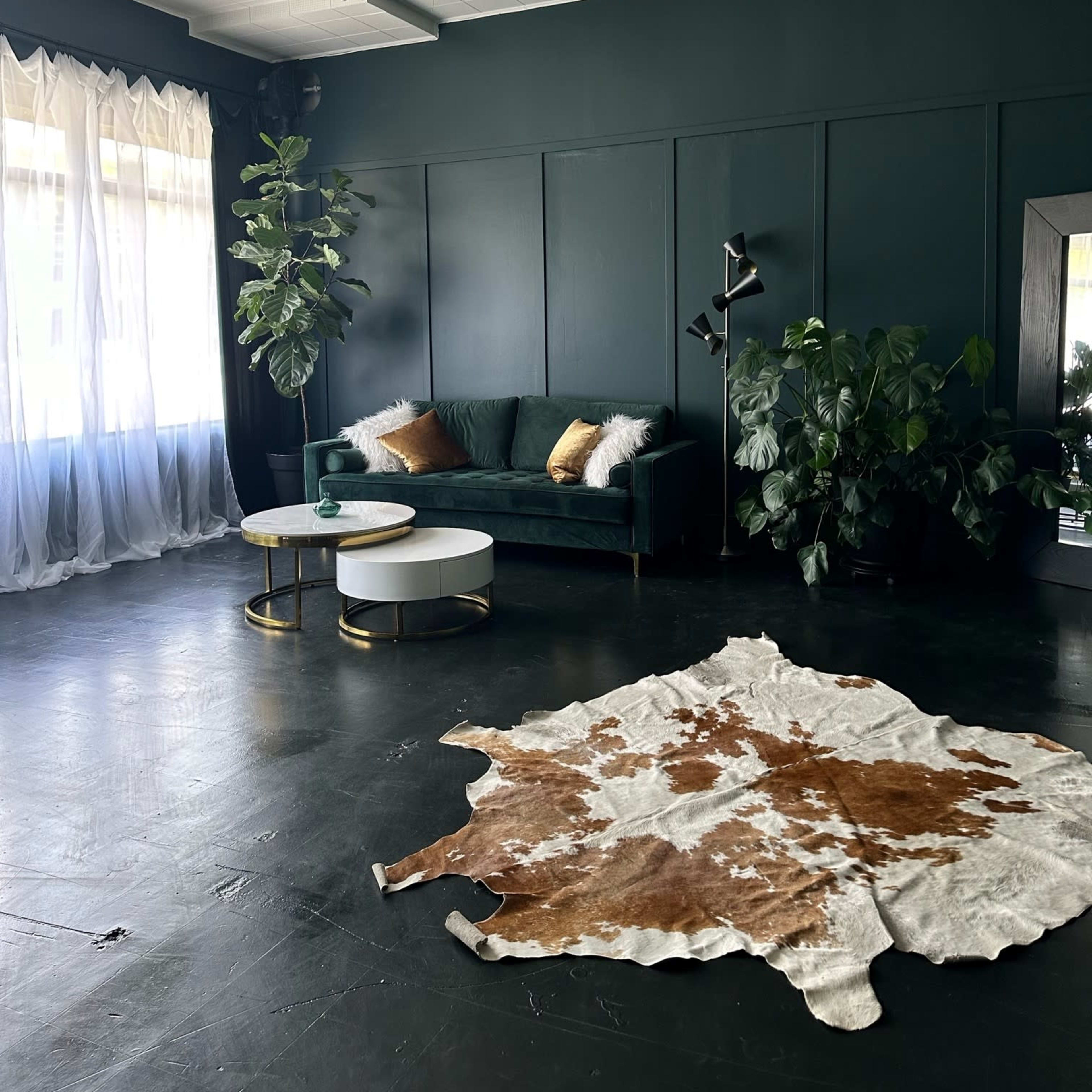 A modern lounge area with a green couch, a round coffee table, a cowhide rug, and plants beside large windows with sheer curtains.