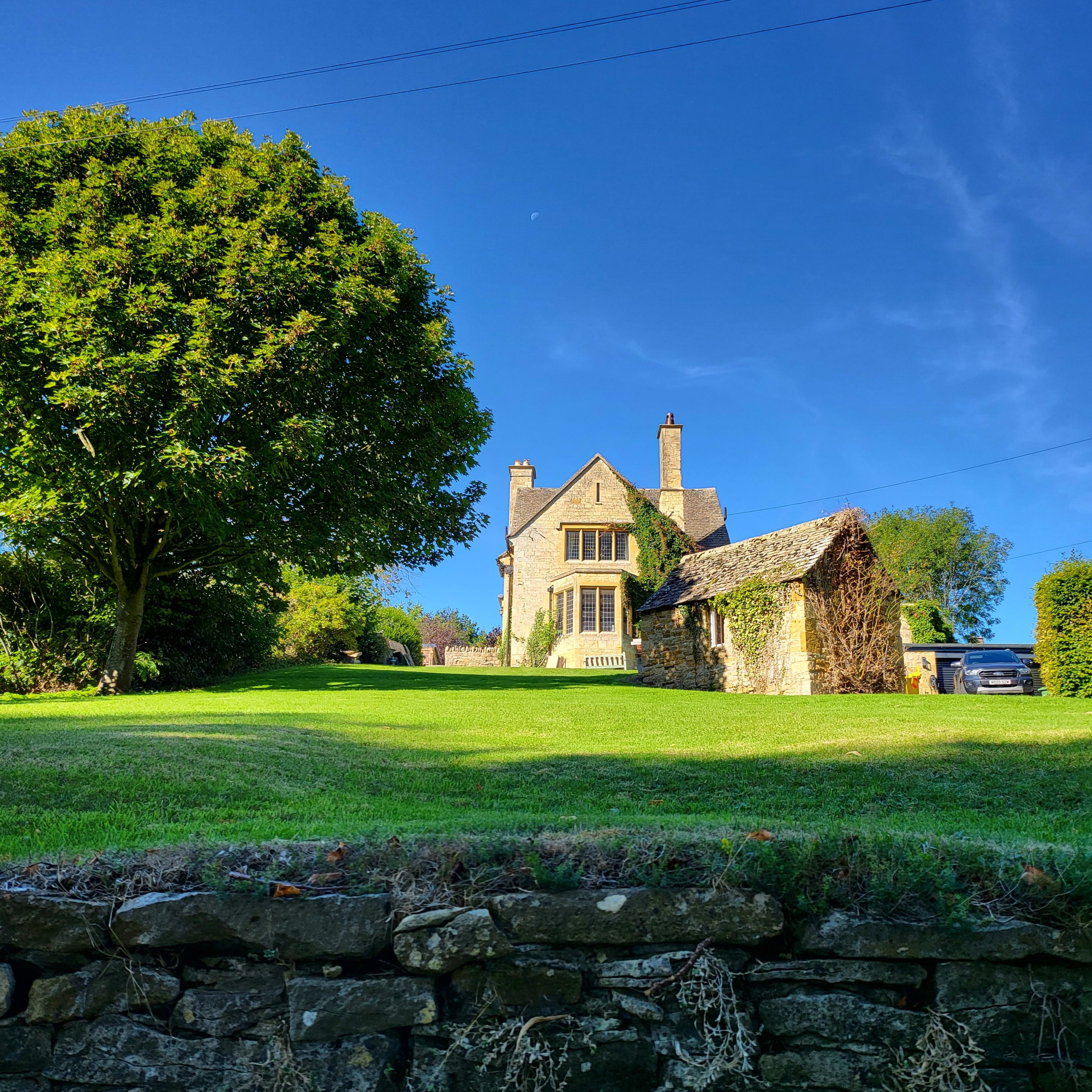 A stone house with tall windows sits on a green hillside, surrounded by trees under a clear blue sky.