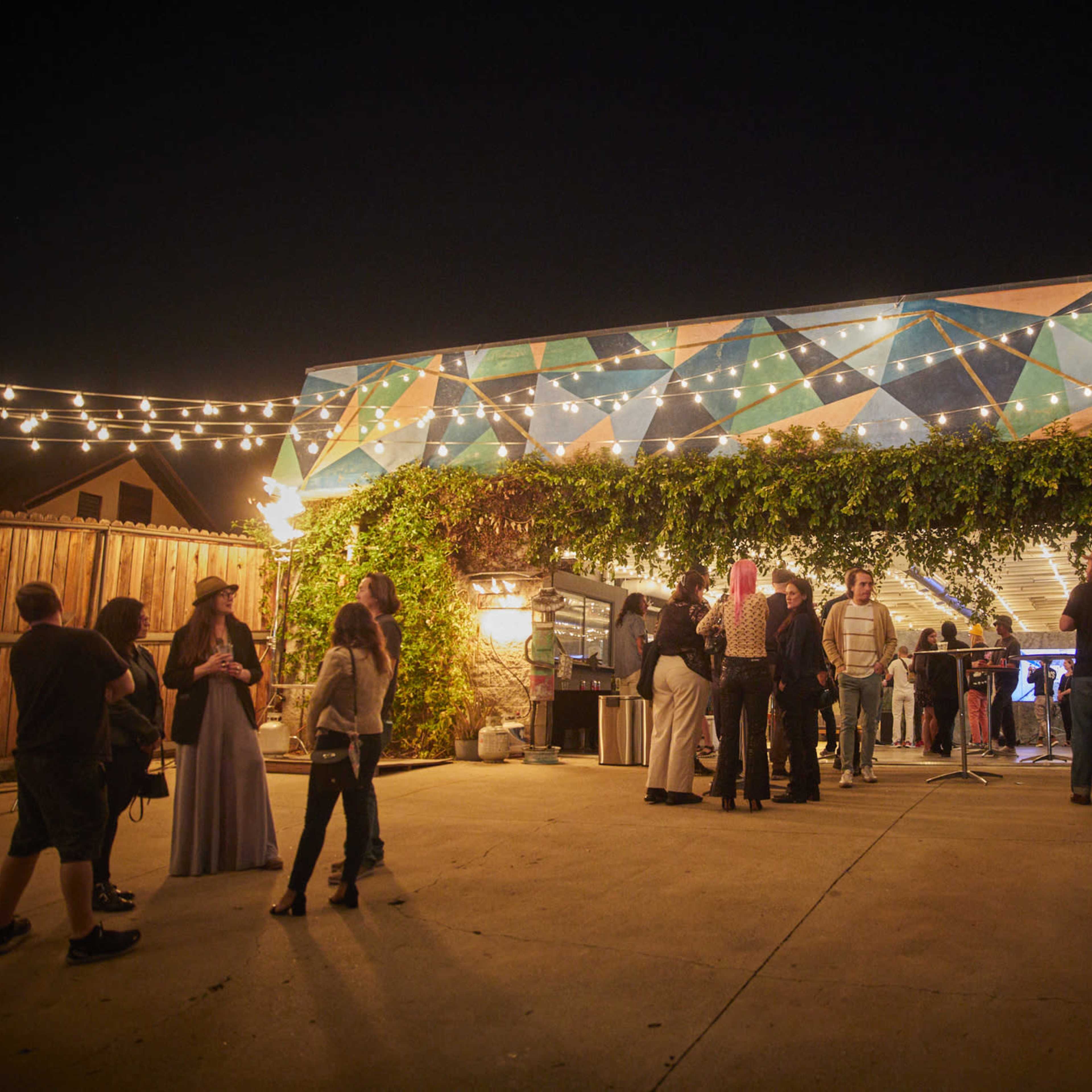 A crowd gathers outside a colorful venue adorned with string lights and greenery during the night.