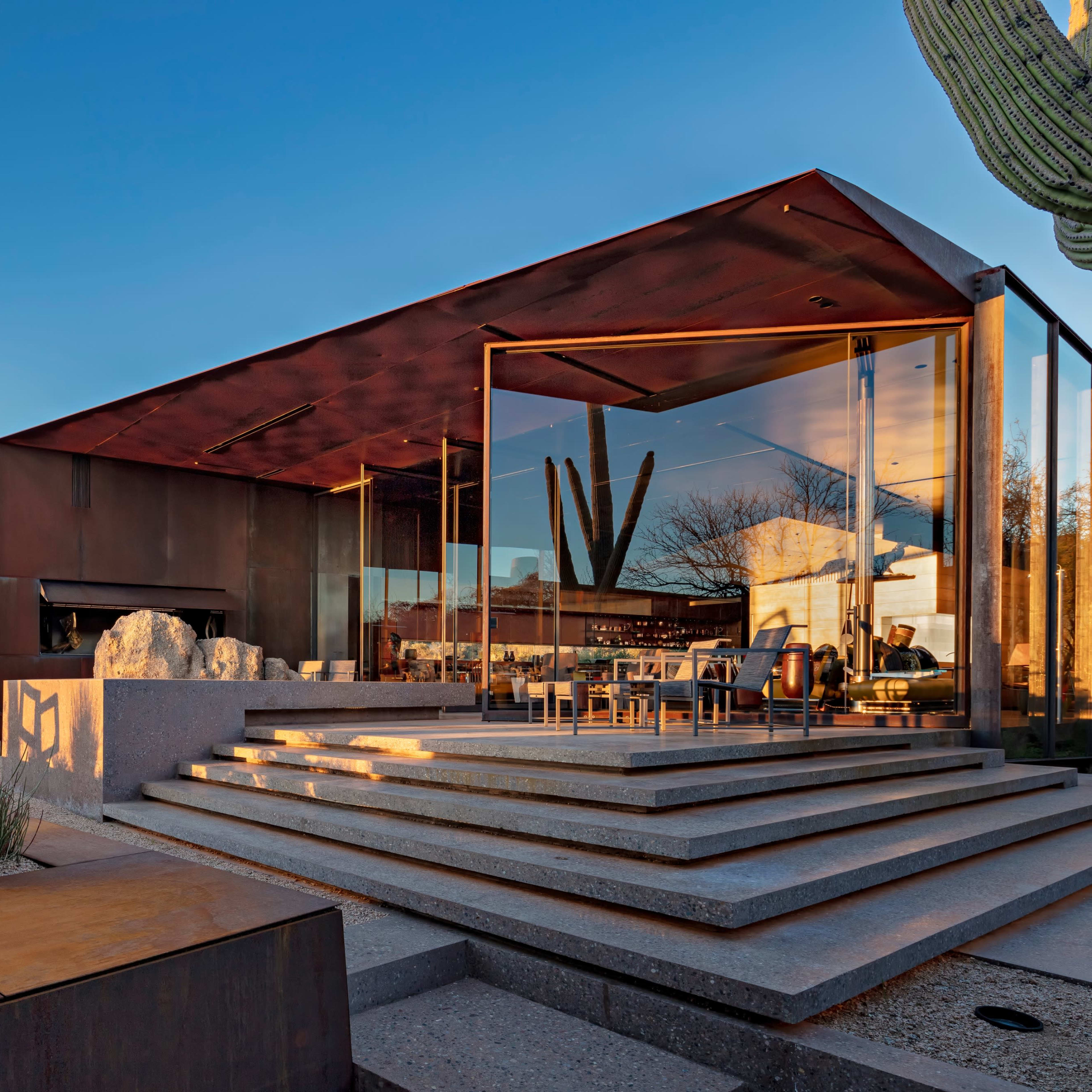 A modern house with a metal exterior and large glass windows is situated in a desert landscape, featuring an outdoor seating area and surrounding cacti.