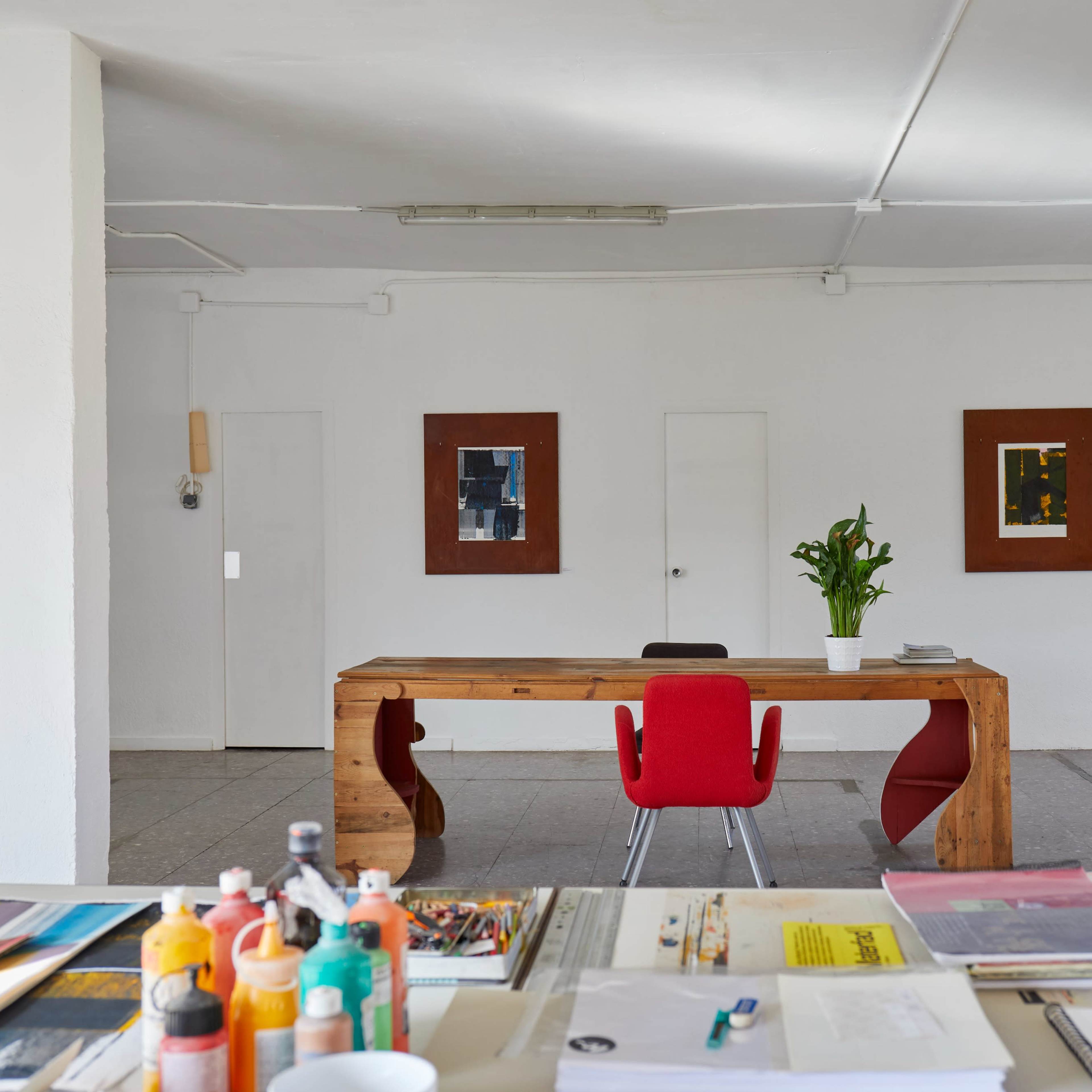 A minimalist office space with a wooden desk, a red chair, and artworks hanging on the walls.