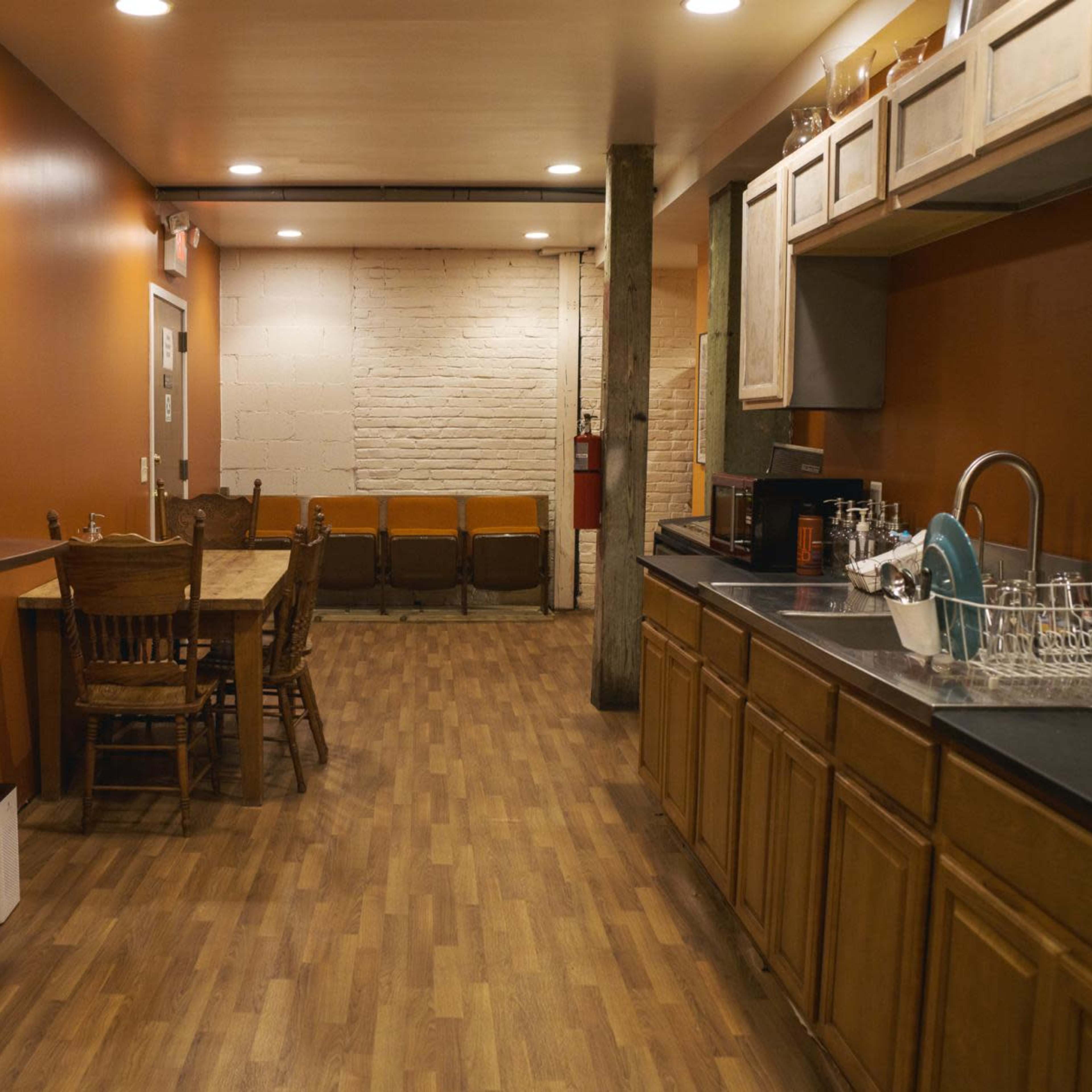 A kitchen area with wooden cabinets, a table with chairs, and a coffee maker against an orange wall, leading into a sitting area with brown chairs.