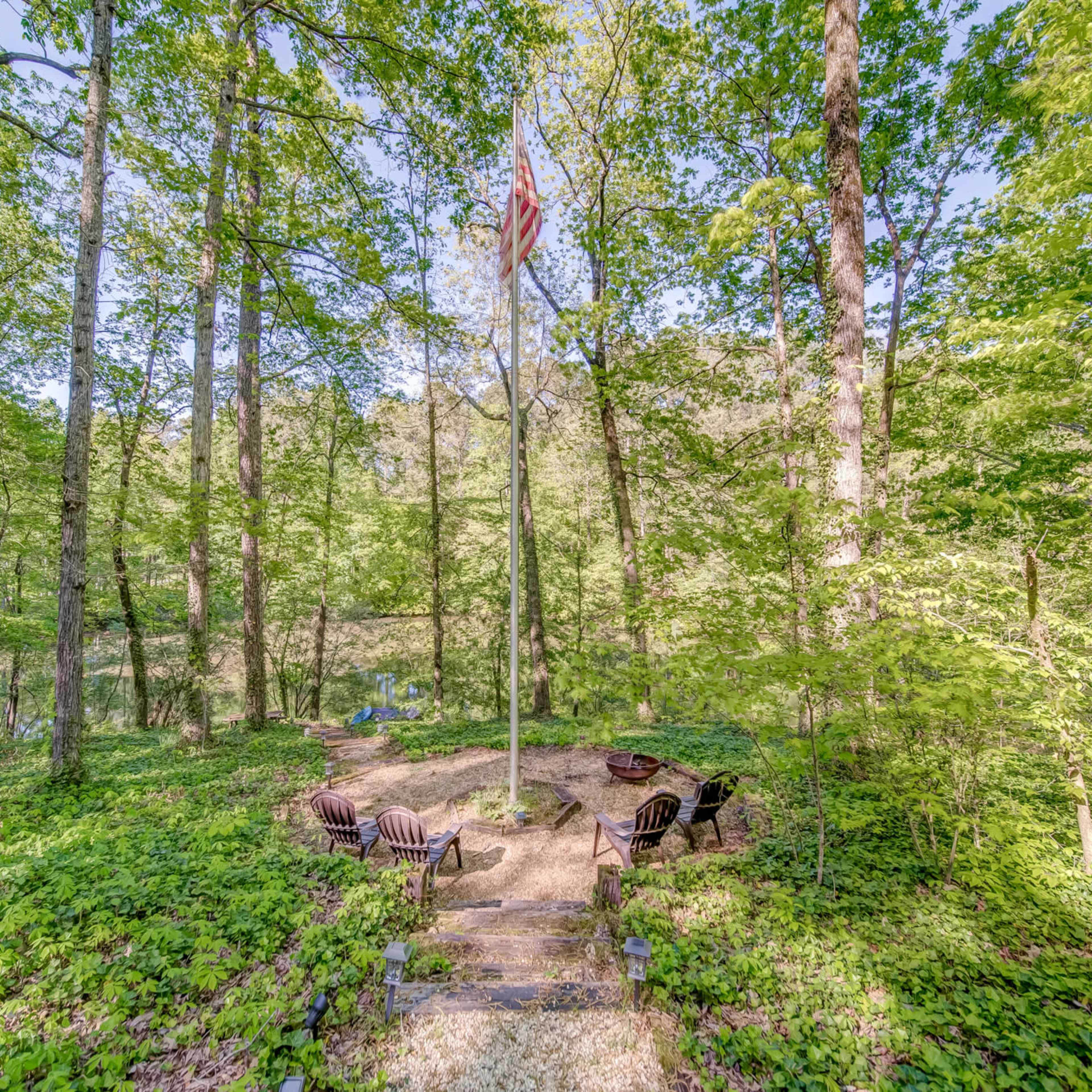 A circular fire pit area with wooden chairs is surrounded by dense green trees in a forested setting.
